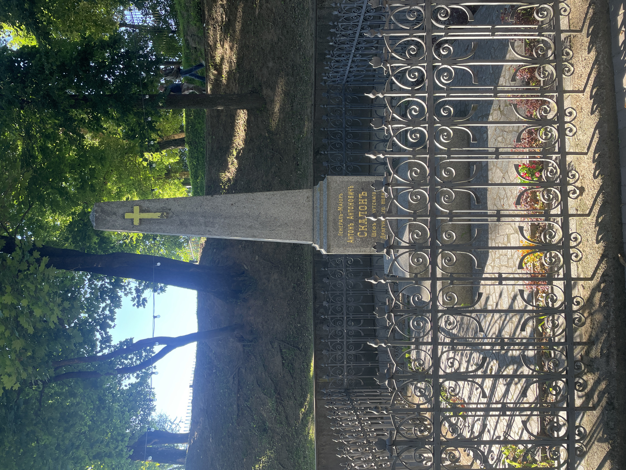 The image depicts a monument in a cemetery located in Sof'ino, Russia. The monument is a tall, grey obelisk with a golden cross at its apex. Inscriptions are visible on the front of the monument. The monument is surrounded by a decorative black metal fence. Beyond the fence, flowers are planted around the base of the monument. The background shows a grassy slope with trees, and two individuals can be seen walking in the distance. The scene is bathed in sunlight, casting shadows on the ground.