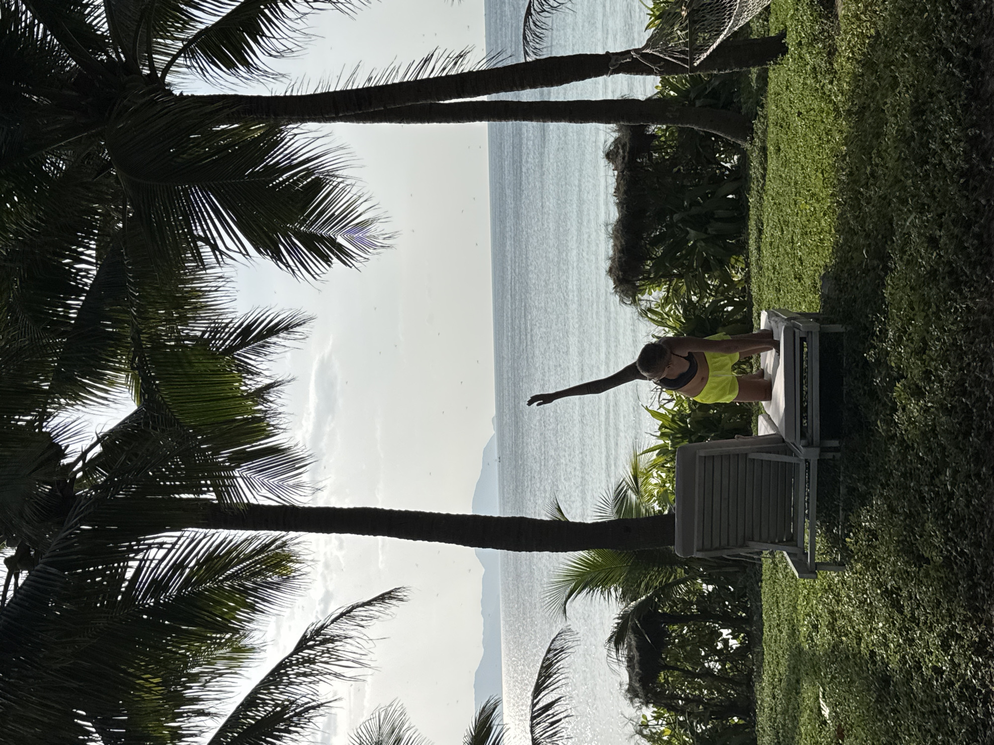A young woman in neon yellow shorts and a black sports bra is performing a yoga pose on a lounge chair on a grassy area. She is on all fours, with one arm extended upwards and her body twisted to the side. Behind her, palm trees frame a view of the ocean and distant islands. A hammock is visible hanging from a tree to the right. The scene is set in a tropical environment. The location is likely a resort or beach area in Đông Ninh Hòa, Vietnam, given the presence of palm trees and the ocean.