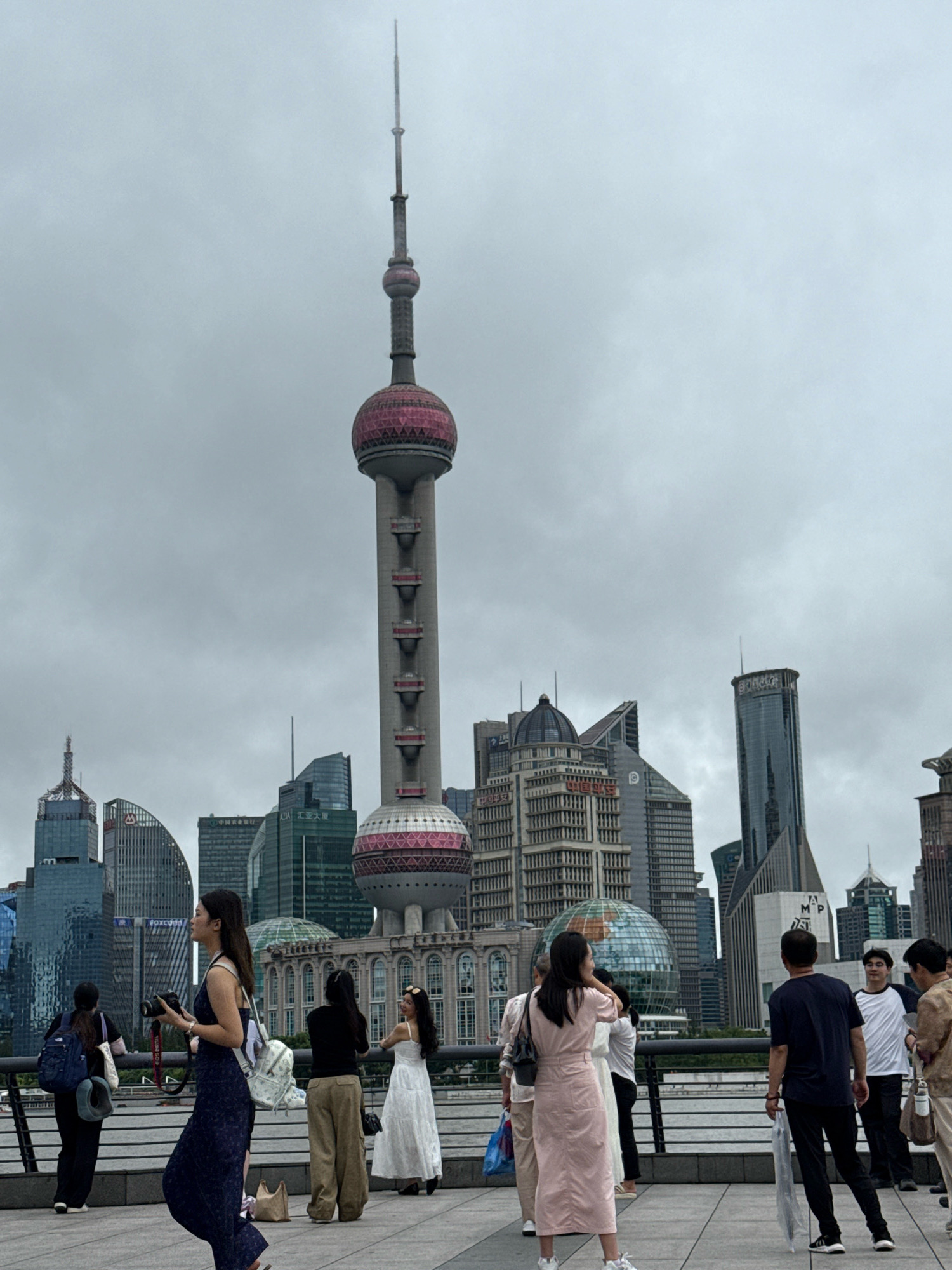 The image depicts the Oriental Pearl Tower, a prominent landmark in the Pudong district of Shanghai, China, against a cloudy sky. In the foreground, a group of people are visible on a promenade, some taking photographs and others observing the cityscape. The individuals are casually dressed, suggesting a leisurely outing. The background is dominated by a collection of modern skyscrapers, characteristic of Shanghai's financial hub. A body of water, likely the Huangpu River, is visible in the lower portion of the image, with a railing separating the promenade from the water. The overall atmosphere appears to be overcast, but the scene is well-lit.