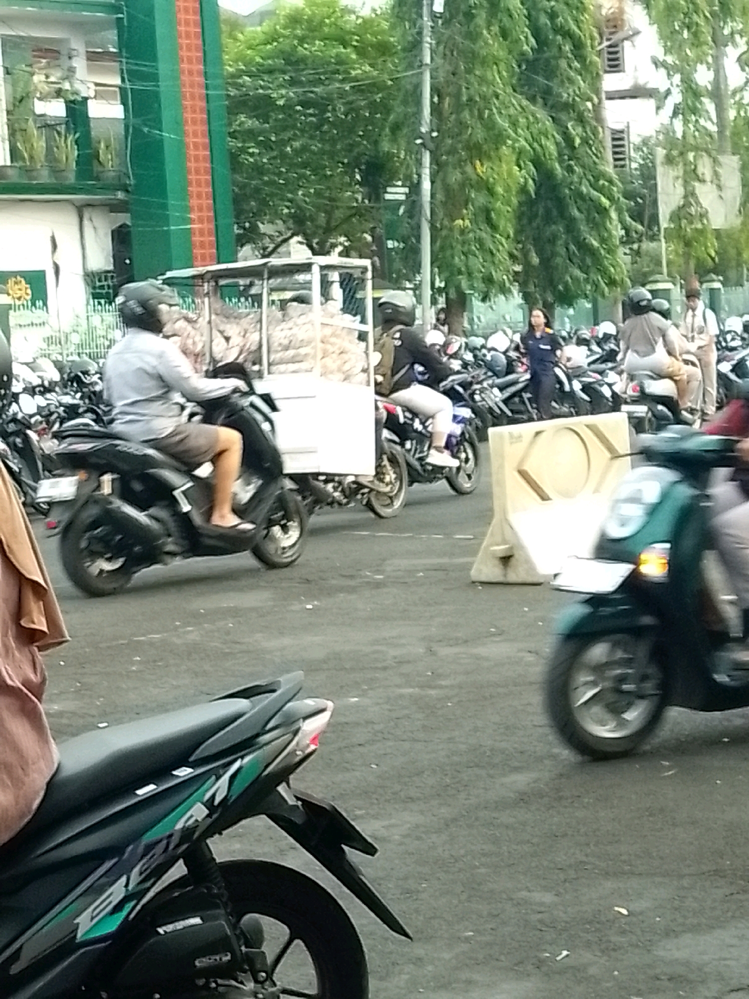 The image depicts a street scene in Jember, Indonesia, characterized by numerous motorcycles and a vehicle carrying goods. In the foreground, a black scooter with green accents is visible from the rear quarter, its rear wheel in motion. To the left, a person in a brown garment is partially visible.
Further into the scene, a person wearing a gray shirt and shorts is riding a black scooter, maneuvering through traffic. The scooter is pulling a small trailer laden with what appears to be produce or goods. Behind this, more motorcycles are parked or in transit, and several people are on foot, including a woman in a dark blue uniform and a person in a school uniform.
The environment suggests an urban setting, possibly a market area or a busy street. A tall building with green and orange vertical stripes is visible on the left, possibly a mosque or public building. Trees with dense foliage line the background, indicating a warm climate. A white traffic barrier is positioned in the middle ground, suggesting roadwork or traffic management. The overall atmosphere is one of moderate activity and daily life in a Indonesian city.