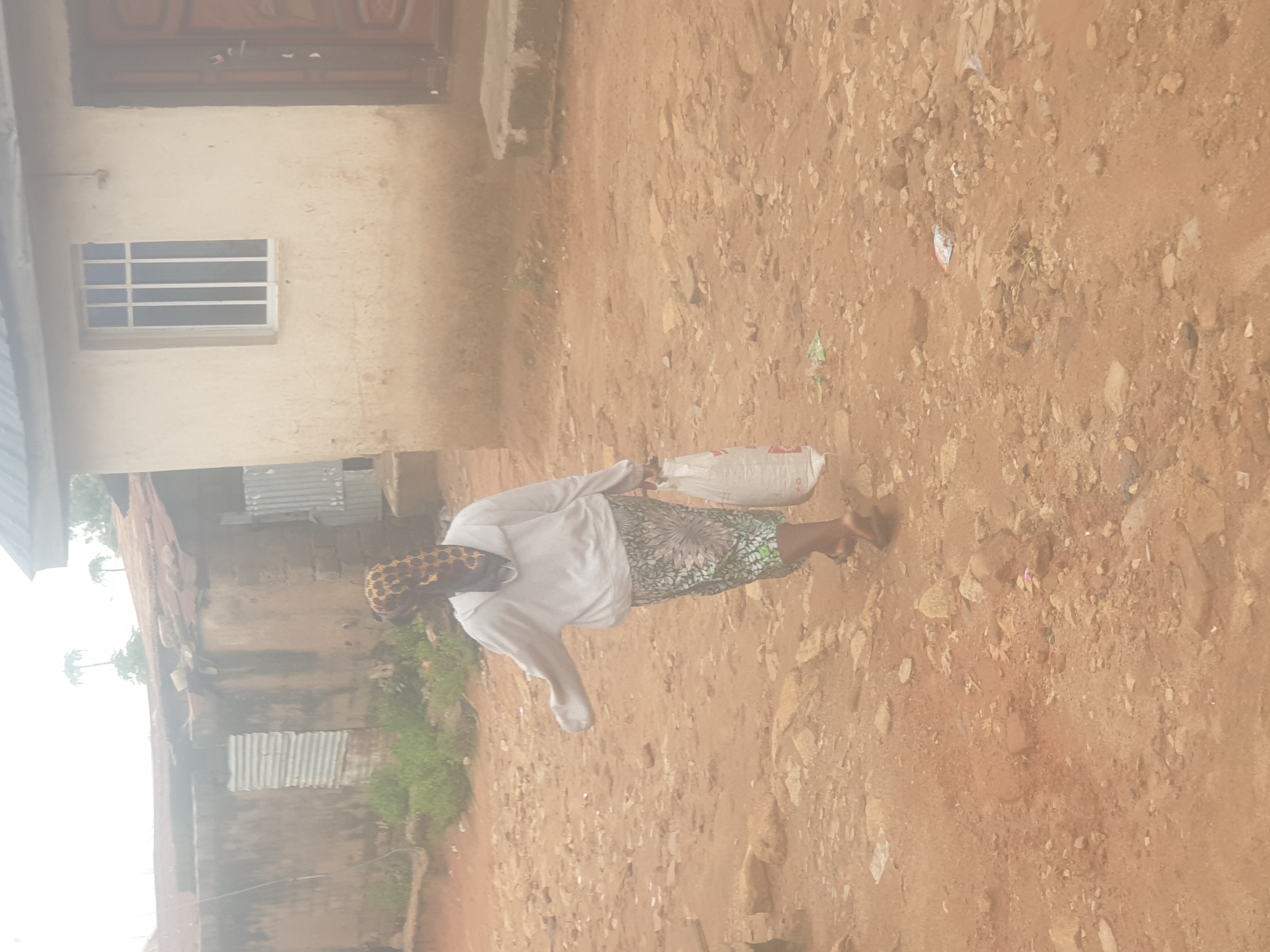 A full shot captures a woman walking on a dirt path, carrying a white plastic bag in her right hand. She is wearing a light-colored hooded sweatshirt, a patterned headscarf with a gold, black, and white leopard print, and a long skirt with a green and white floral pattern. Her feet are clad in dark sandals. The ground is uneven, covered with dirt and scattered stones. In the background, a light-colored building with a dark door and a white-paned window is visible. To the left, another building with a corrugated metal section and a rusted tin roof is present. The scene appears to be an outdoor environment in a residential area. The image is not located in Washington, United States of America.