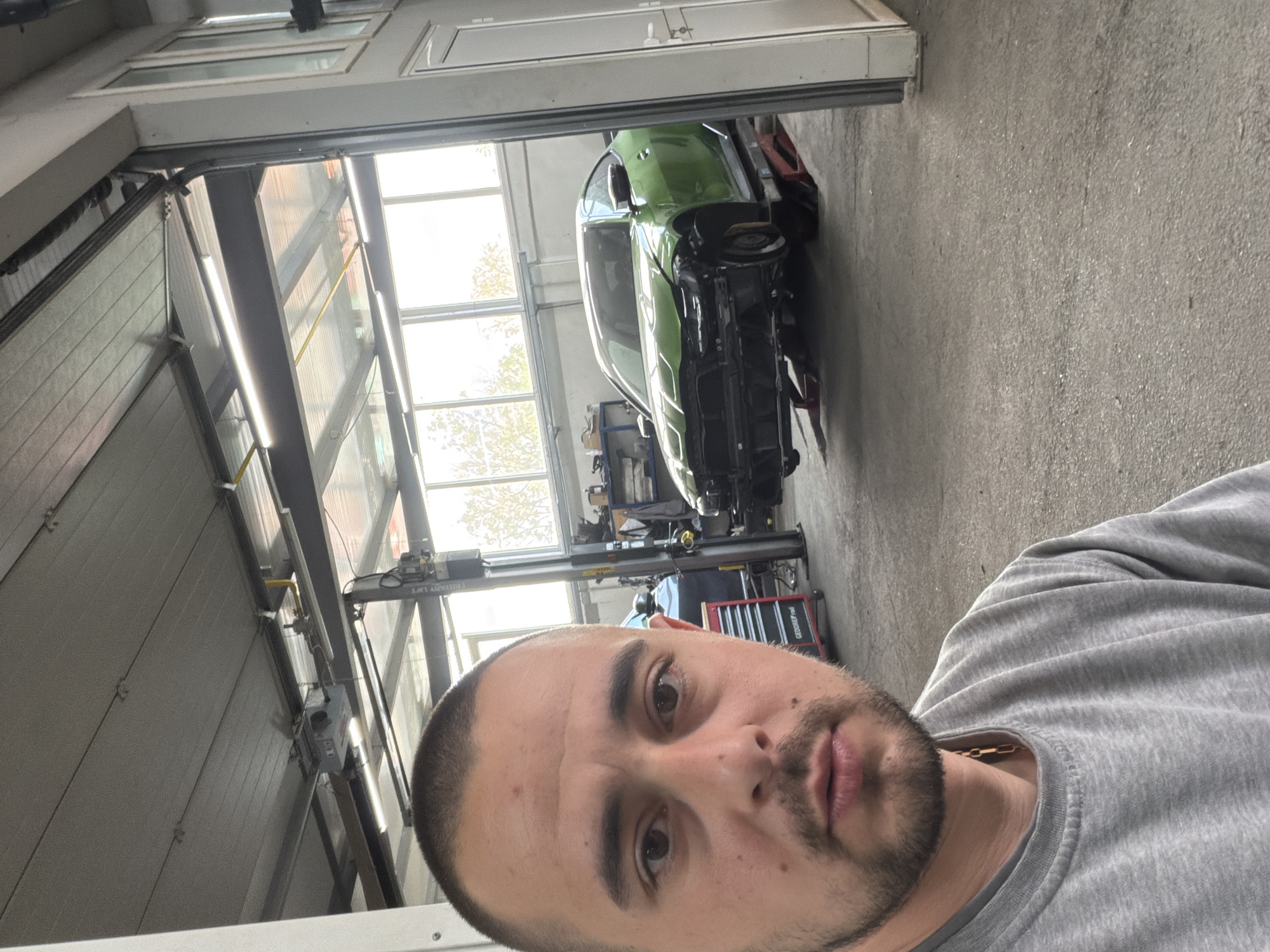 A man is pictured in what appears to be a garage or auto repair shop. The man is facing the camera, wearing a grey shirt, and has short hair and a beard. Behind him, a green car is visible, partially disassembled on a lift. The garage has a high ceiling with bright lighting and large windows. Tools and equipment are scattered around the area. The garage door is partially open. It is not possible to determine the specific location in Burgas, Bulgaria, based solely on the provided image.