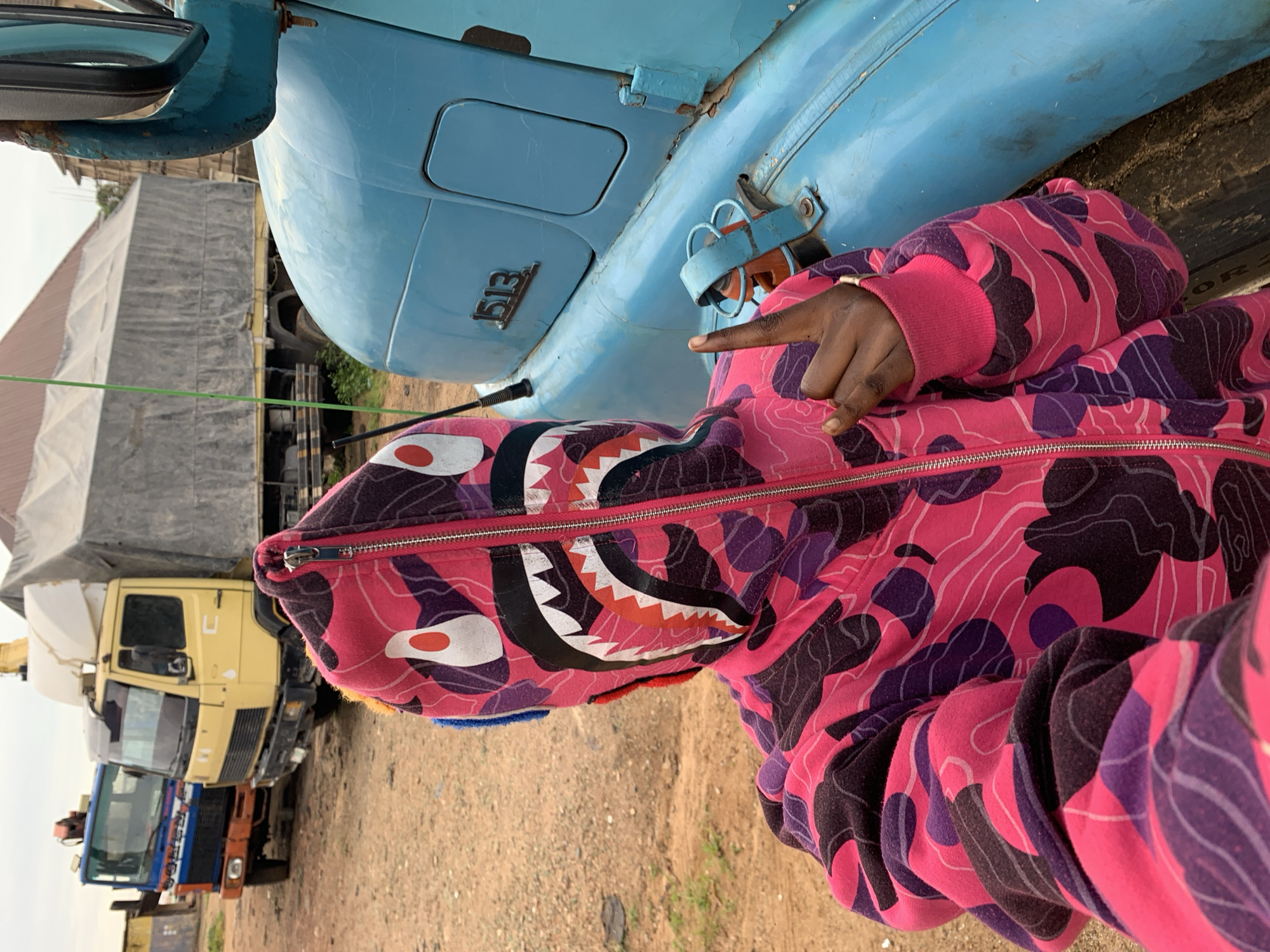 A person is depicted in a full-body, hooded sweatshirt featuring a pink, purple, black, and white camouflage pattern with a shark face design on the hood, obscuring the wearer's face. The sweatshirt is zipped completely, and the person's right hand is raised, with the index finger extended upward and the other fingers curled. The scene is an outdoor, unpaved area with reddish-brown dirt and sparse green vegetation. In the midground, to the right of the person, is the light blue cab of a truck, featuring the embossed numbers "1513" on its side and an antenna. In the background, two other trucks are visible: a yellow Mercedes-Benz-style truck with a tarp-covered cargo bed and a folded crane, and further left, a blue truck with an orange bumper. Portions of buildings or structures with reddish-brown roofs are visible in the far background under an overcast sky. The image is set in Rafin Par, Nigeria.