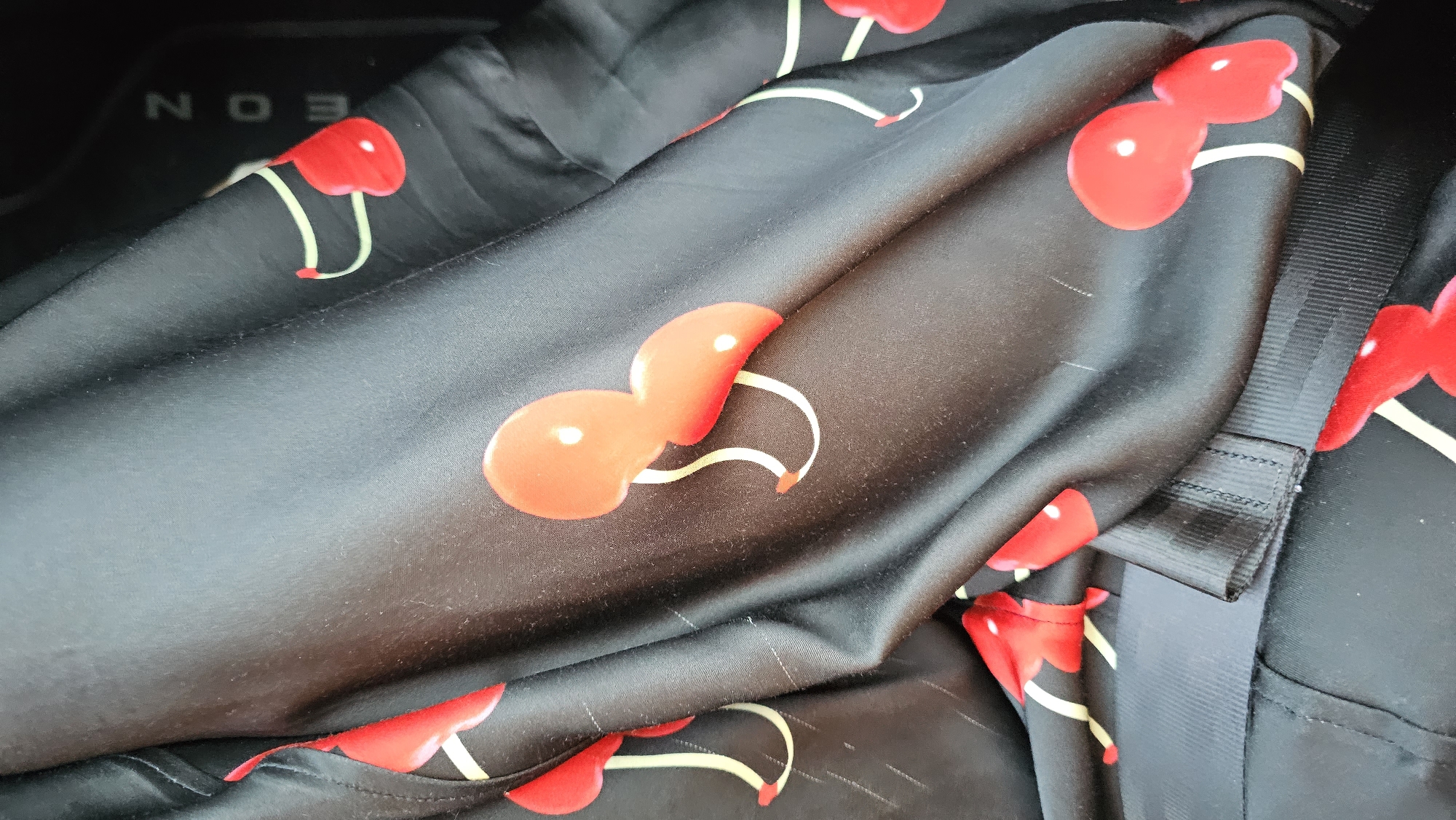 A close-up shot of black fabric with a cherry print. The fabric is draped and creased, with some light reflections on the surface. There are several stylized red cherries with yellow stems scattered across the fabric. In the background, the letters "EON" are visible, vertically oriented and slightly out of focus, on a dark surface that could be part of a car interior. A black strap runs horizontally across the bottom of the image, partially obscuring the fabric. The scene appears to be indoors, possibly in a car, in Les Marines, Spain.