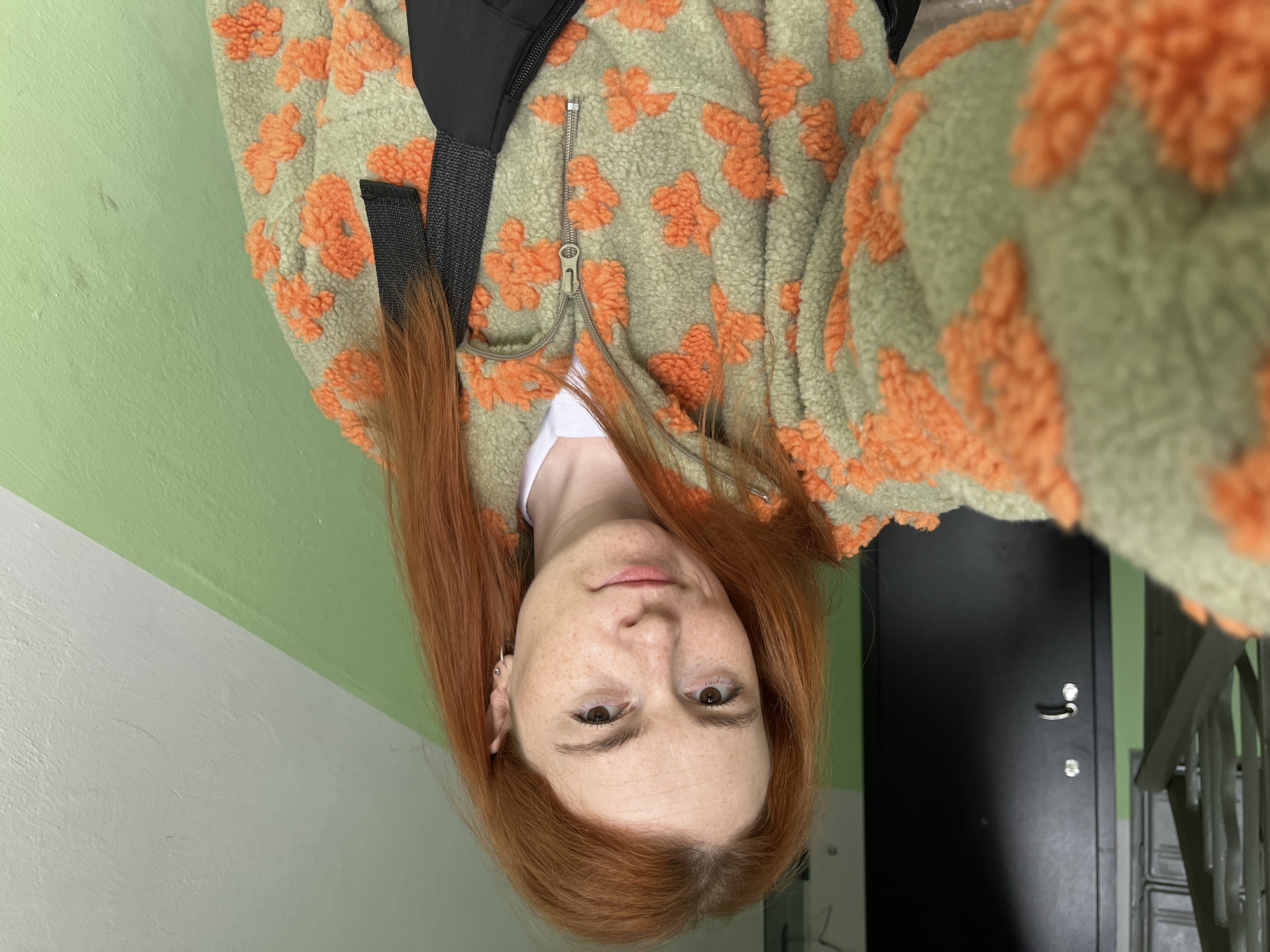 This is a selfie taken by a young woman with long, reddish-orange hair and fair skin with freckles. She is wearing a green fleece jacket with an orange floral pattern and a white t-shirt underneath. The image is oriented sideways, so the woman's head is towards the bottom of the frame.
She is looking directly at the camera with a neutral expression. Her eyes are brown. She has a small hoop earring in her left ear. A black bag strap is visible over her shoulder.
The background appears to be an indoor hallway. To the left, there is a light green wall. To the right, there is a dark gray door with a silver handle and lock. A metal railing is visible on the far right, suggesting a staircase. The lighting is consistent with an indoor environment, possibly daytime, as there are no strong shadows or artificial light sources that are immediately obvious.
There is no visible text in the image. The overall impression is a casual, personal photograph taken in what seems to be a residential building or common area.