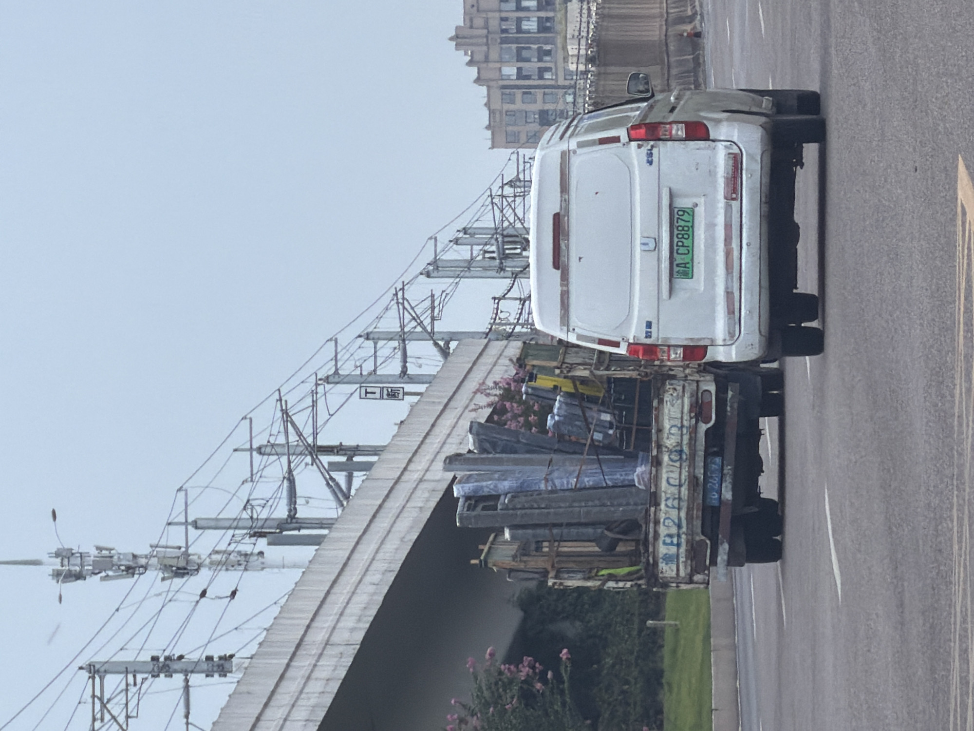 A white van with license plate "A CP8879" is driving on a highway. To its left, a truck is also driving on the highway, carrying a load of various long, flat objects, including what appears to be building materials or large furniture. The truck's license plate is partially visible and reads "2G 26575". The vehicles are driving under an elevated overpass, with power lines and communication towers visible above. In the background, apartment buildings are visible. The sky is overcast. The scene appears to be in a city in China, specifically Jiangjin.