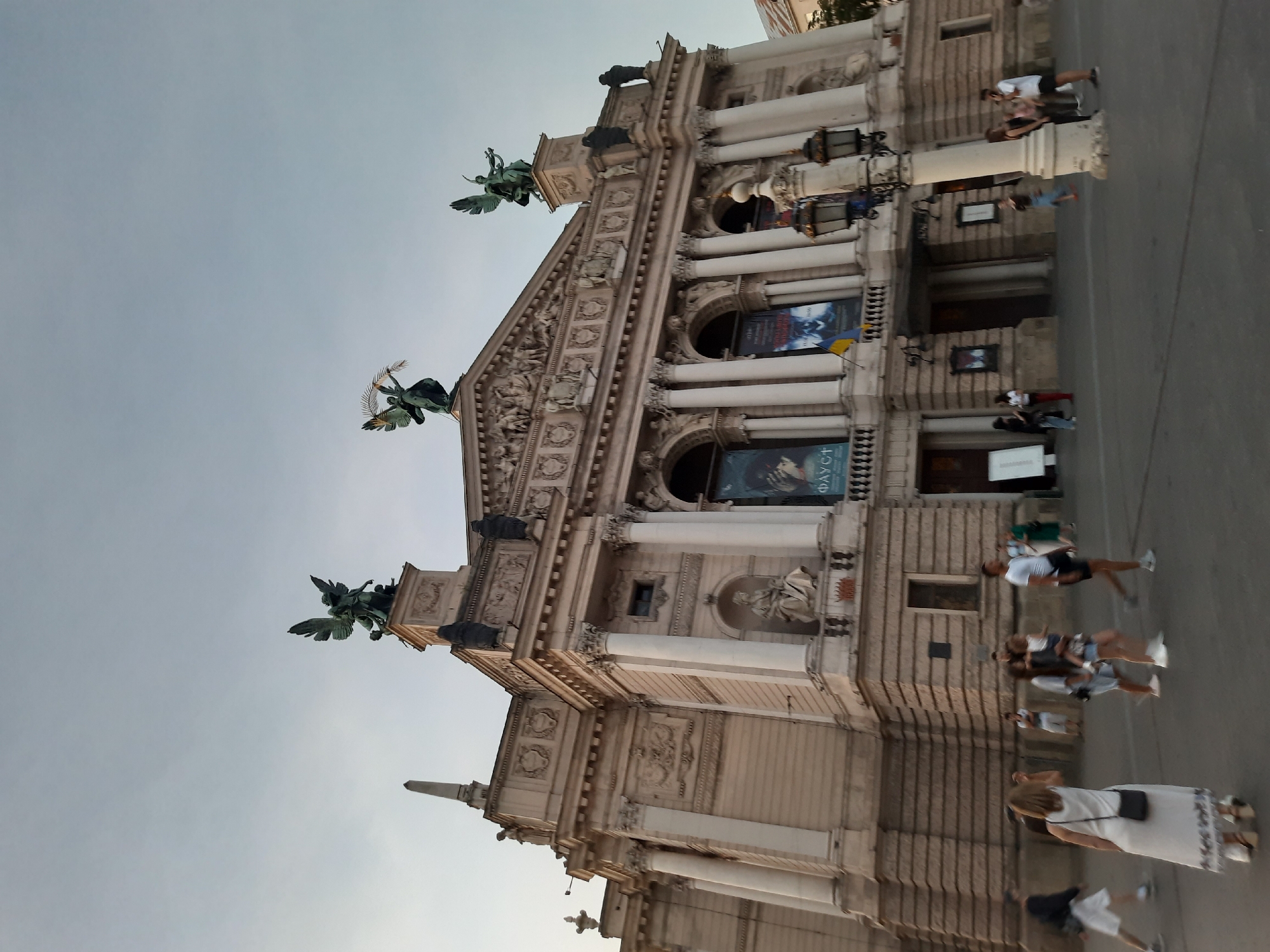 This is a low-angle, outdoor, medium shot of the Lviv Opera House in Lviv, Ukraine. The sky is overcast and pale blue. The grand facade of the Opera House dominates the frame, featuring classical architectural elements such as columns, pediments, and ornate sculptures. Atop the building, several bronze statues of winged figures are visible, including one holding a laurel wreath. Banners hang from the building's facade, with text in a foreign language. A Ukrainian flag is also displayed. Several people are walking in the square in front of the Opera House. In the foreground, a woman in a white dress is walking away from the camera. To her left, a man in shorts and a dark shirt walks past. Other individuals and small groups are scattered throughout the scene. A decorative lamppost stands in the right foreground. The ground is paved.