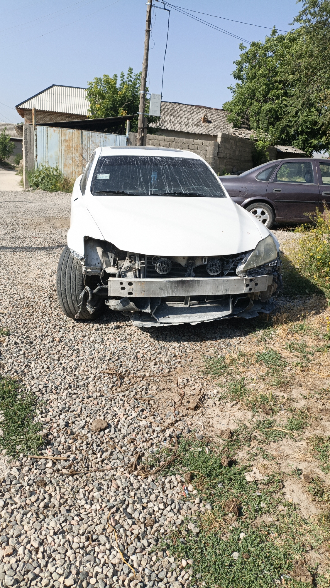 A damaged white sedan is parked on a gravel lot. The front of the car is heavily damaged, with the bumper missing and the headlights exposed. A dark-colored sedan is parked behind it. The scene appears to be in a residential area with houses and trees in the background. The sky is clear and blue. The image was taken on a sunny day.