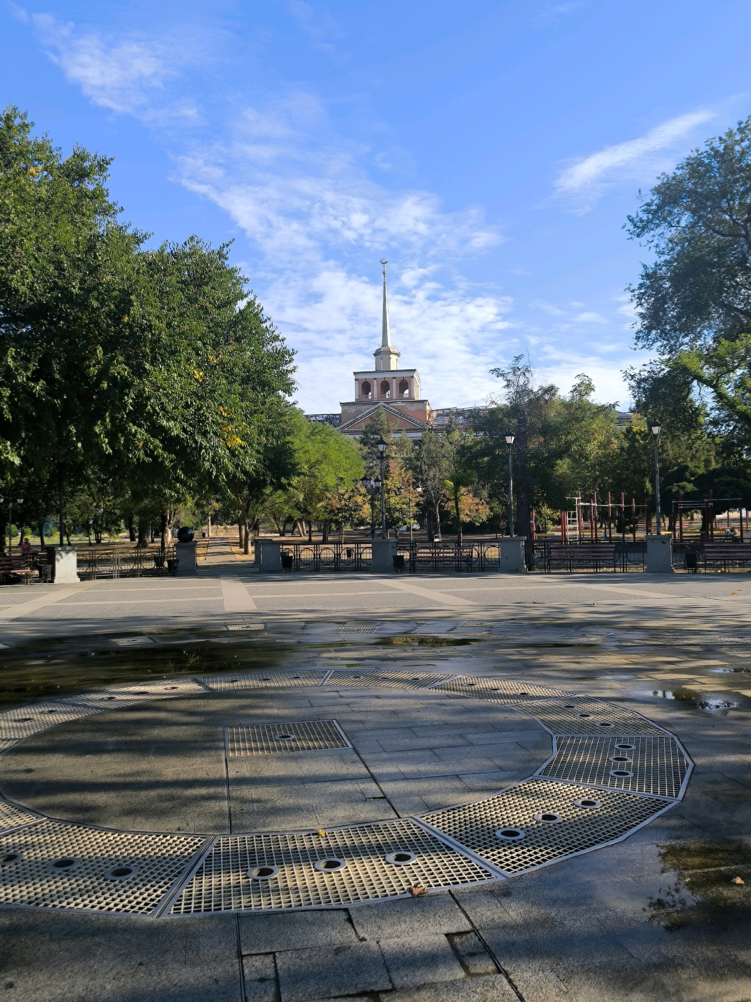 The image shows a paved public area with a circular grate system in the foreground, possibly part of a dry fountain. Beyond this, a park with trees, benches, and lampposts is visible. In the background, a multi-story building with a spire stands against a bright blue sky with scattered white clouds. This scene is characteristic of a public square or park, and the building's architecture suggests it could be a historical or governmental structure within the city of Mykolaiv, Ukraine. There are no people clearly visible in the foreground, but a few figures can be discerned in the background within the park area. The lighting suggests it is daytime, with sunlight casting shadows on the pavement.