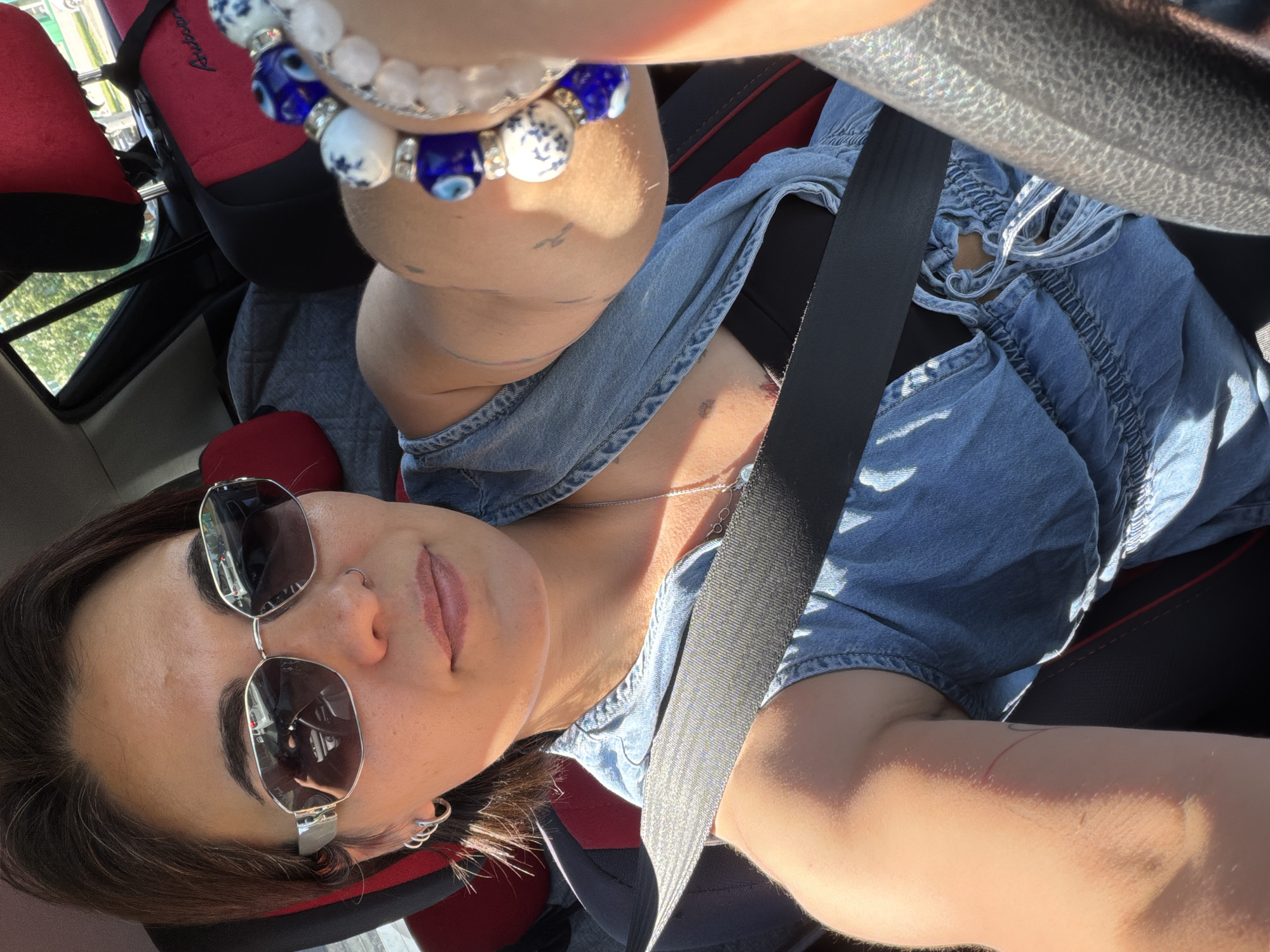 A medium shot captures a woman in the driver's seat of a car, taking a selfie. She is wearing sunglasses, a denim jumpsuit, and a black undershirt. A seatbelt is fastened across her chest. On her right wrist, she is wearing several bracelets, including one with blue and white beads. The interior of the car is visible, including red and black upholstery on the headrests and seats. The background shows trees and a bright, sunny sky, indicating an outdoor setting. The image was taken in the city of Petropavlovsk-Kamchatsky, Russia.