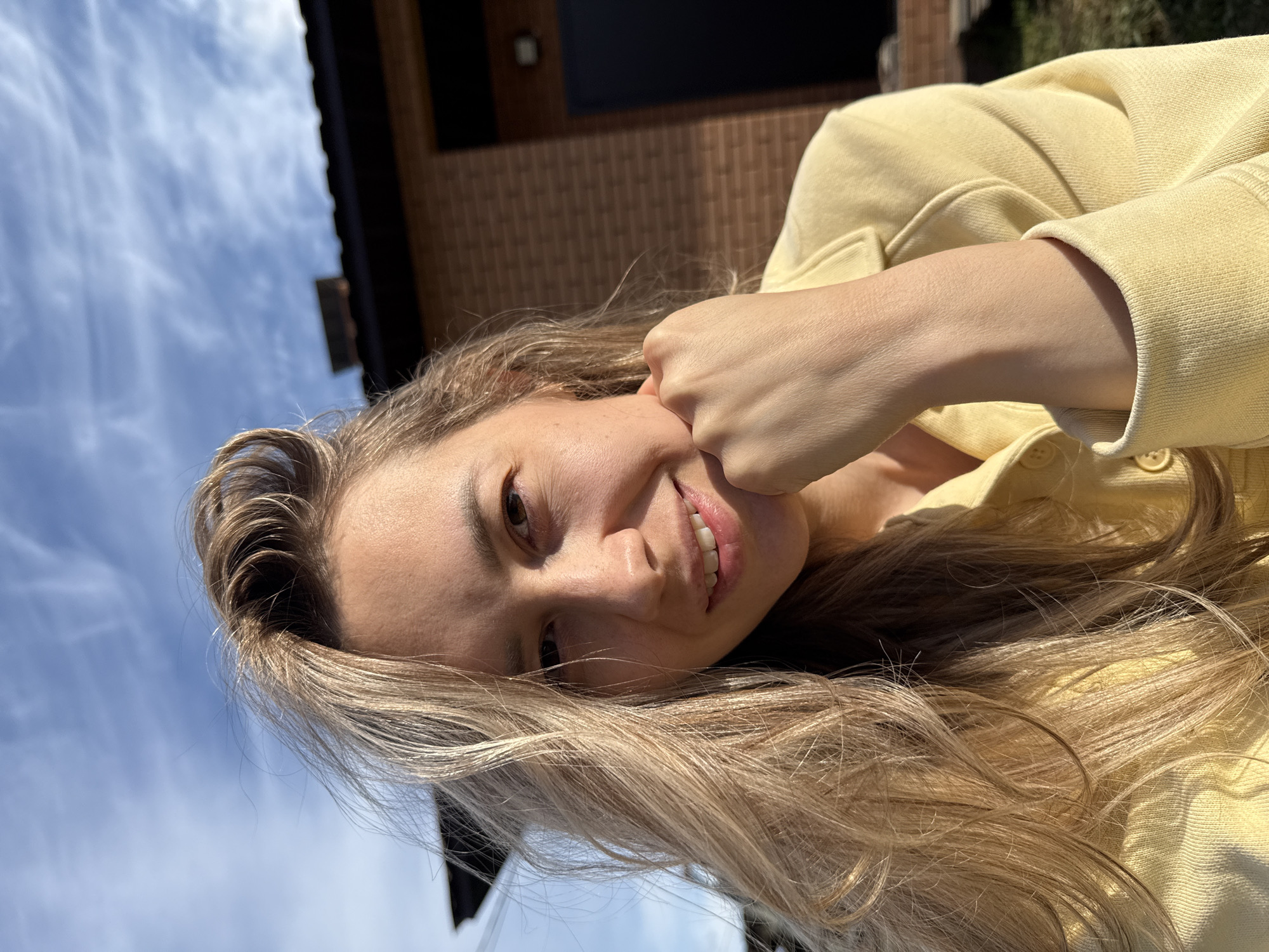 A smiling woman with long blonde hair, wearing a yellow top, is positioned in front of a brick building under a partly cloudy sky. Her left fist is near her face. The location appears to be in an outdoor setting in Chaikovsky, Russia.