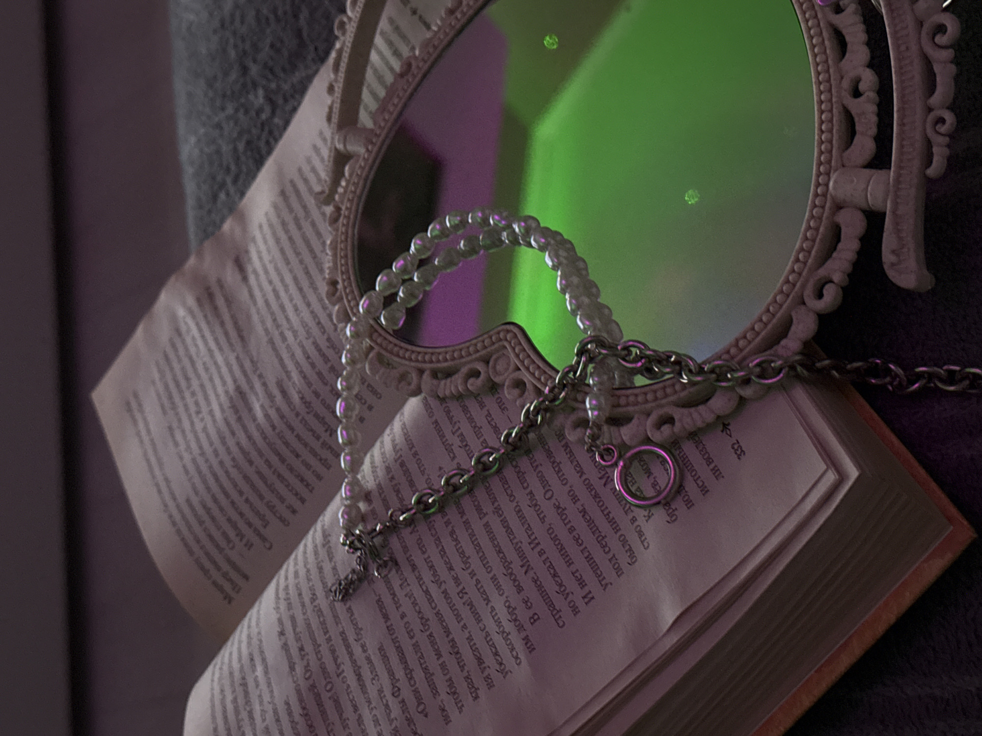 A close-up shot shows an open book, a pearl necklace, and a silver chain placed on top of it, with a vintage-style mirror reflecting green and purple light in the background. The book's pages are filled with dense text in a foreign language. The pearl necklace drapes across the book and the edge of the mirror, while the silver chain also rests on the book, with a small ring attached. The mirror, decorated with ornate carvings, reflects a blurry and colorful background. The entire scene is lit with an ambient light, highlighting the textures of the book's pages, the sheen of the pearls, and the luster of the chain.