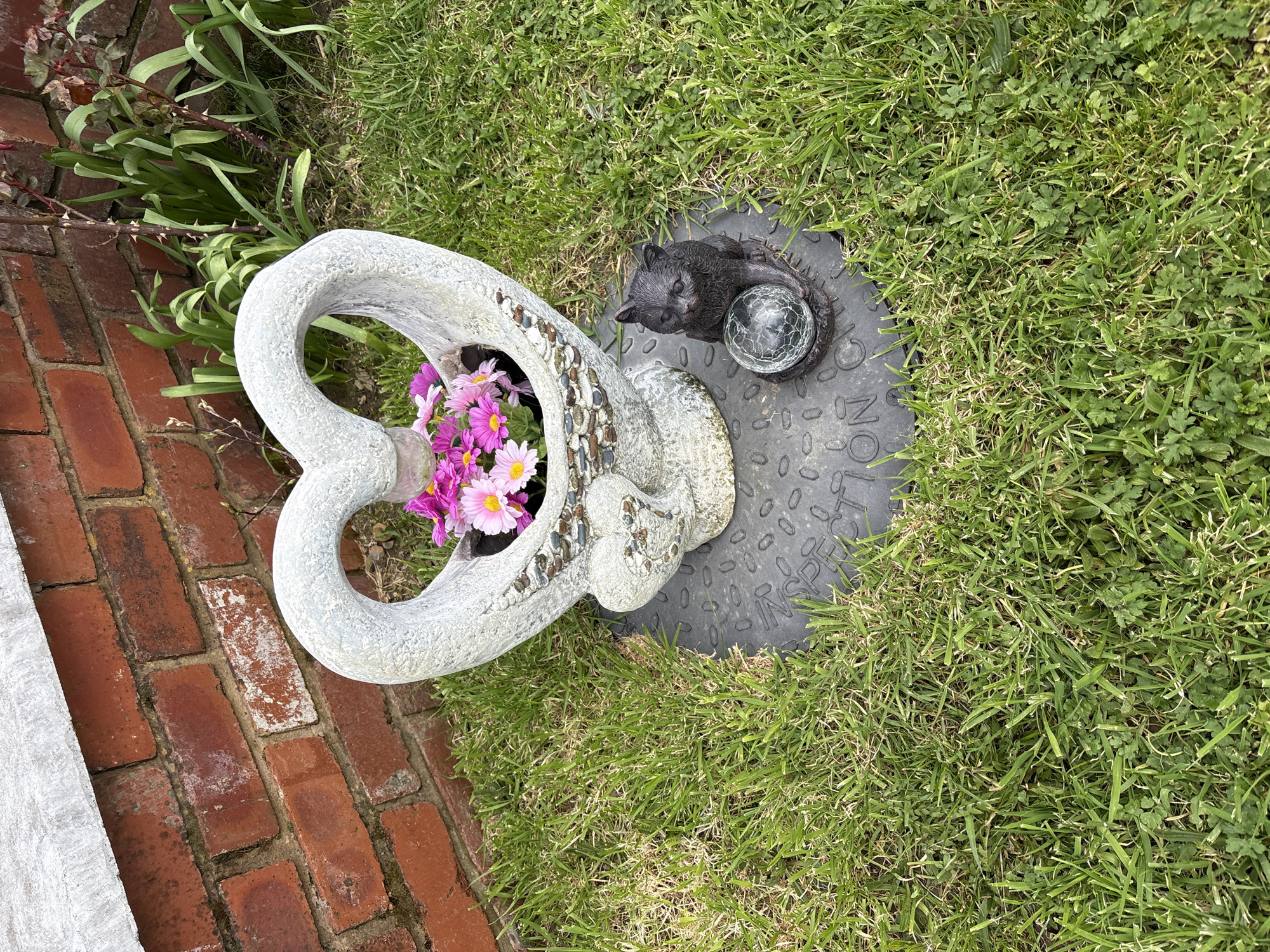 The image shows a memorial display on a patch of grass. A white, heart-shaped stone structure filled with pink and white flowers sits partially on a brick path. Next to it, on a circular gray manhole cover, is a dark gray cat statue holding a clear glass ball. The background is green grass. Location is unknown, and there is no evidence to determine that the location is in Shrewsbury, United Kingdom.