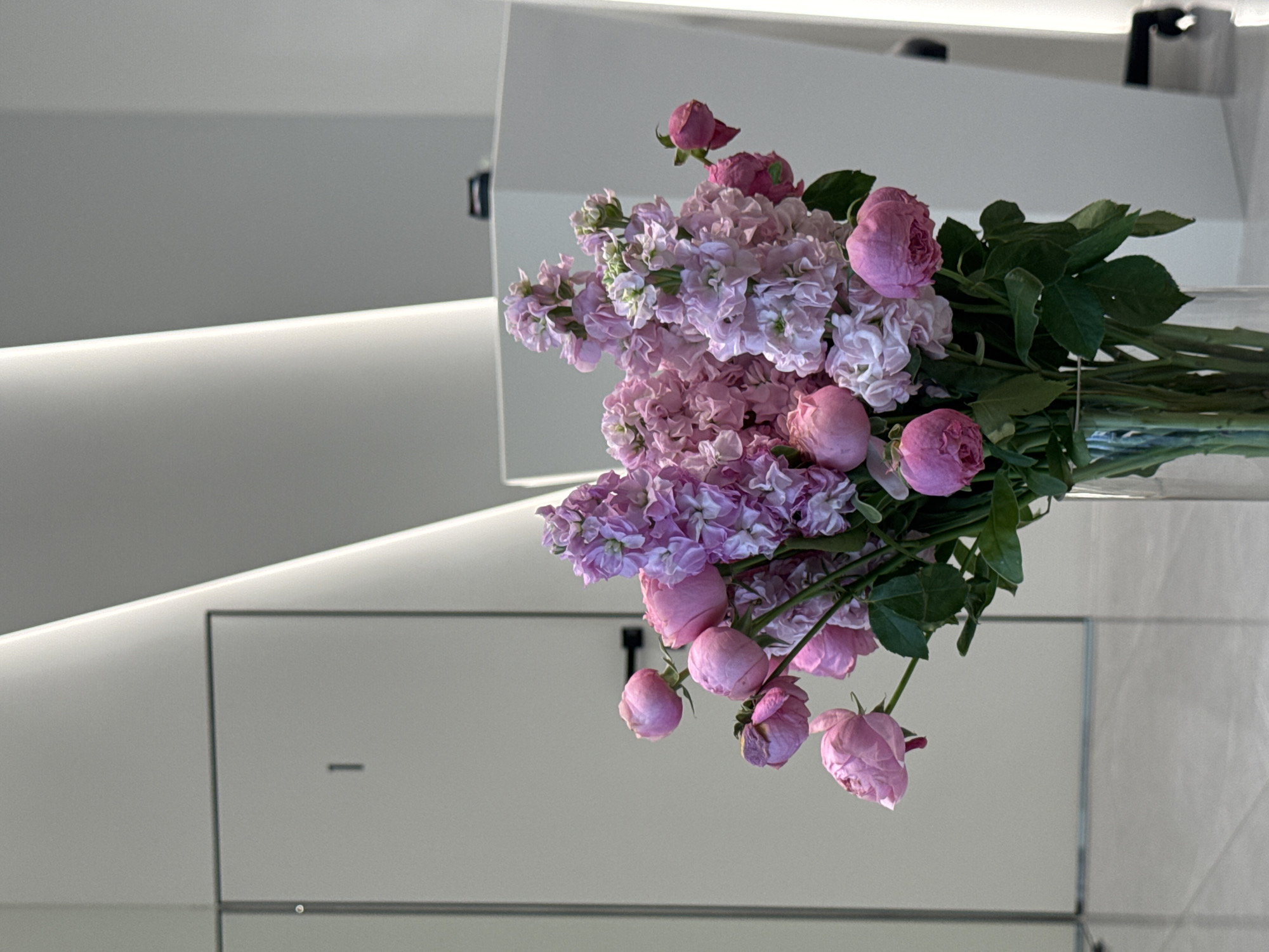 A bouquet of pink flowers, likely roses and stock, is arranged in a clear glass vase. The backdrop is a modern, minimalist interior with a white color scheme and geometric design elements. The location is not verifiable as Saint Petersburg, Russia, as the image does not contain any location specific information.