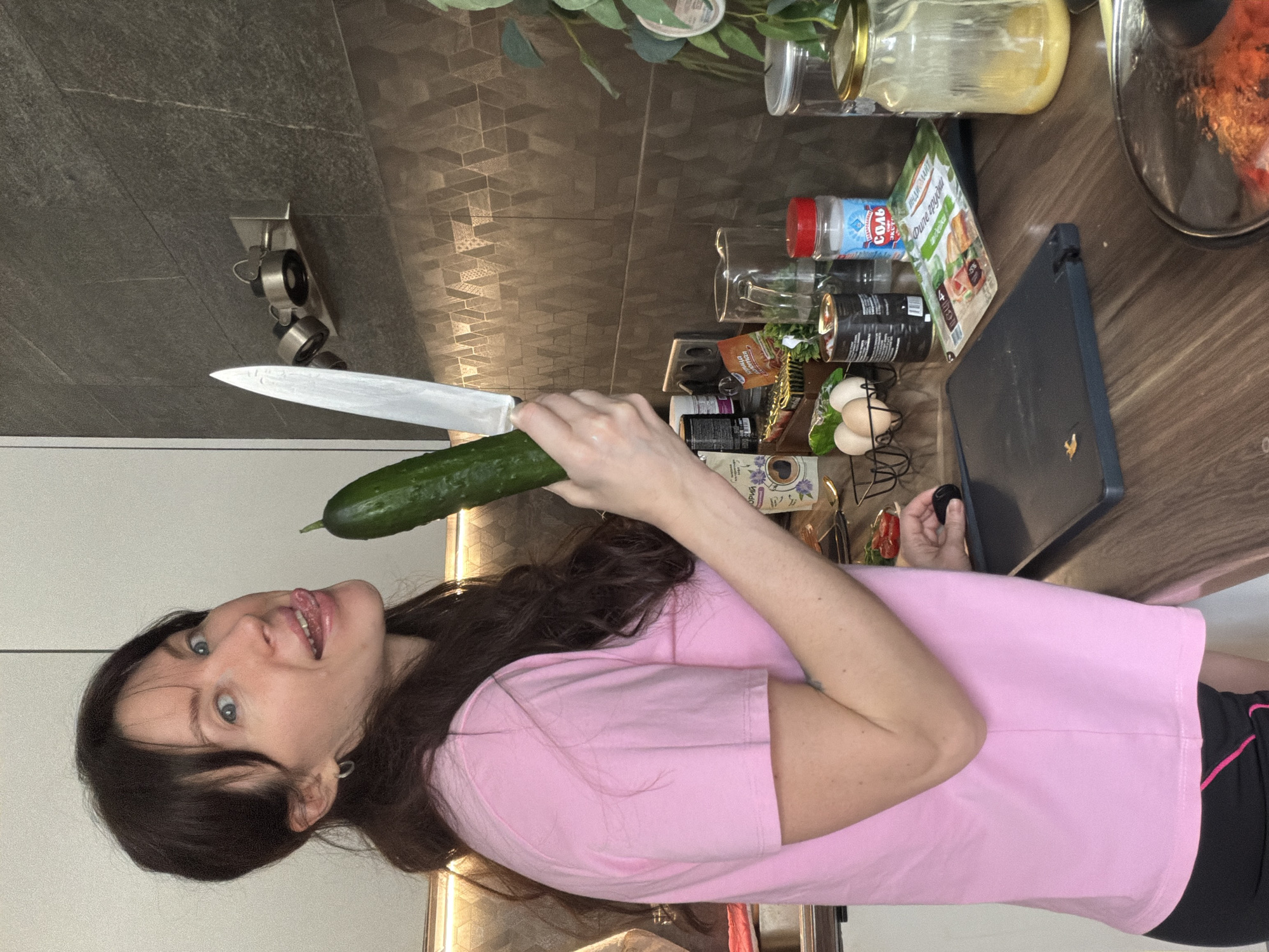 A female subject stands in a modern kitchen in Perm, Russia, looking directly at the camera with a playful expression, her tongue extended. She has dark hair and wears a pink t-shirt over dark bottoms with a pink stripe. In her left hand, she holds a green cucumber, and in her right, a silver-bladed kitchen knife, both raised. The kitchen features a dark brown countertop populated with various food items and utensils, including a black chopping board, a wire basket containing three light-colored eggs, cans (one bearing the Russian text "Филе грудки" from the brand "ИНДИЛАЙТ"), a clear plastic salt shaker labeled "СОЛЬ" in Russian, a clear glass pitcher, and a large lidded glass jar containing a yellowish substance. A small white plate with sliced red items is also on the counter, alongside a potted green plant. A pan with a clear glass lid, containing reddish food, is partially visible at the lower right. The backsplash consists of dark, geometrically patterned tiles illuminated by under-cabinet lighting. A wall-mounted fixture with three spotlights is visible above the backsplash. White kitchen cabinets are present on the left.
