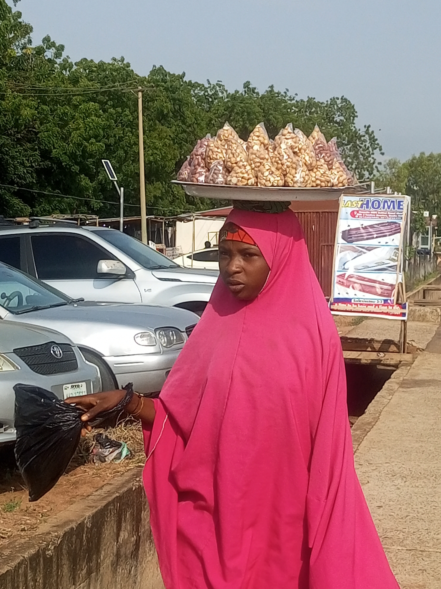 In Gombe, Nigeria, a woman wearing a vibrant pink hijab walks outdoors, carrying a tray of packaged nuts on her head. Her right hand holds a black plastic bag. In the background, several vehicles, including a silver SUV and a silver sedan, are parked. A sign advertising "LASTHOME" coffins stands to the right of the woman, displaying images of different coffin designs. The environment suggests a commercial area with trees and a paved walkway.