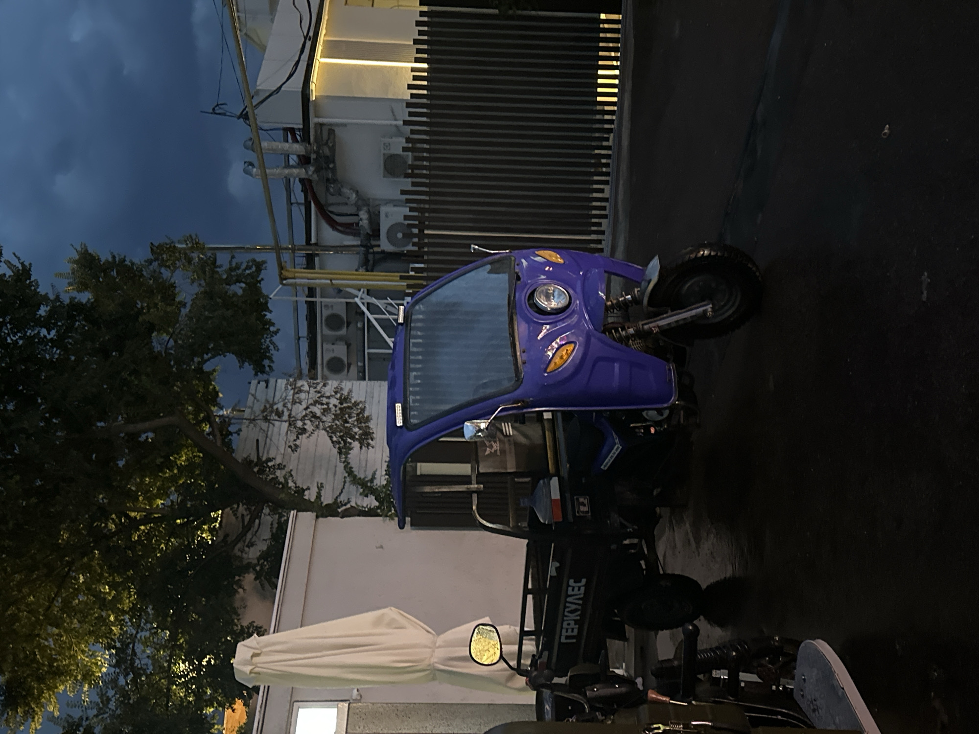 The image shows a purple three-wheeled vehicle parked on a wet asphalt surface in front of a dark wooden fence. The vehicle has a cab with a windshield and a cargo bed. On the side of the cargo bed, the word "ГЕРКУЛЕС" is written in white letters. To the left of the vehicle is a large, cream-colored umbrella, and behind it is a white building. Further in the background, there is a tree with green leaves, and a darker, overcast sky. The scene appears to be outdoors, possibly in a courtyard or street area. The overall lighting suggests it is either dawn, dusk, or a heavily overcast day. There are no people visible in the image. The presence of the name "ГЕРКУЛЕС" (Hercules) in Cyrillic script on the vehicle suggests a potential connection to Ukraine. Given the context of Odesa, this vehicle might be used for local delivery or transport within the city.