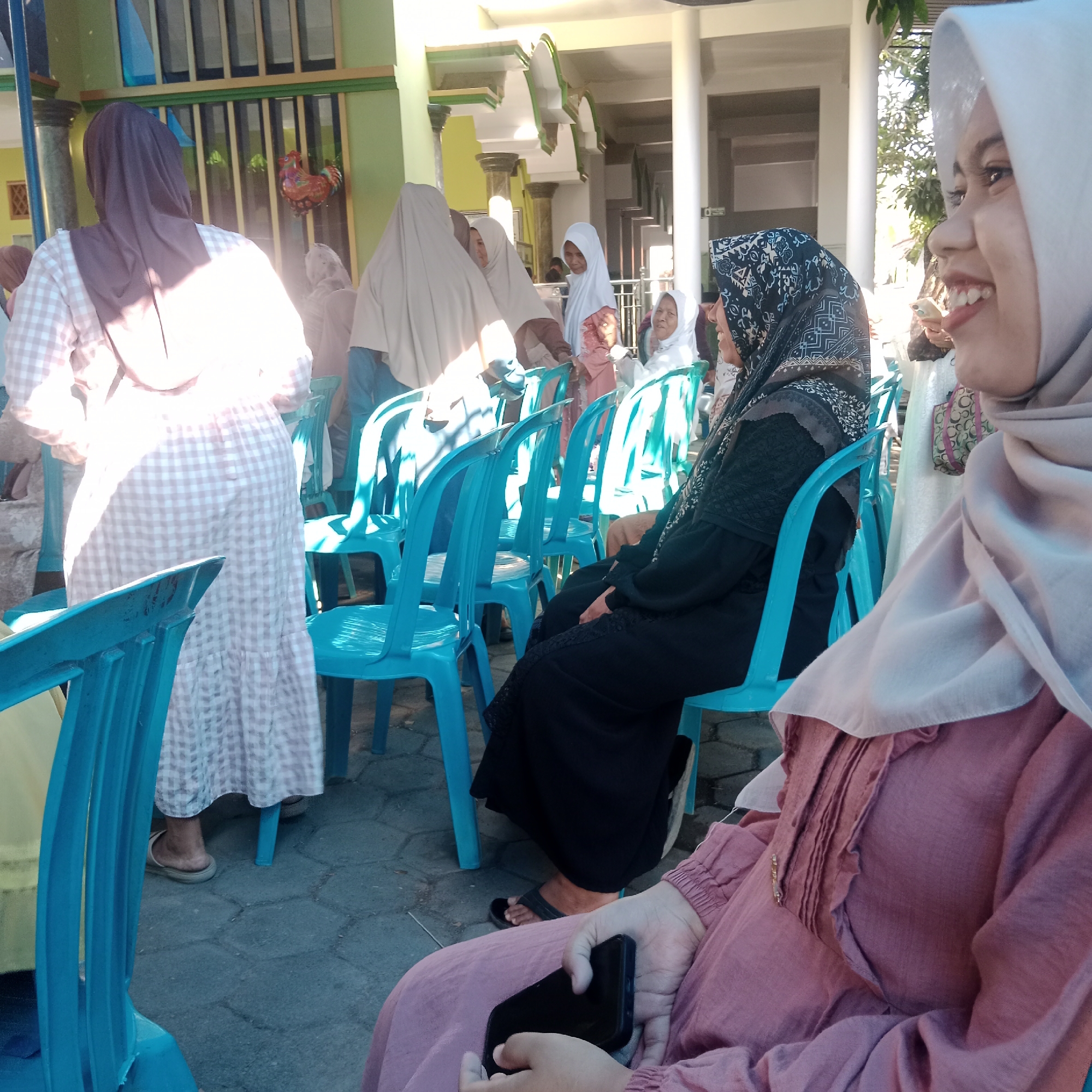 The image depicts an outdoor gathering with several individuals, most of whom are women wearing headscarves. The setting appears to be a community event or celebration in Tegowanu Kulon, Indonesia. In the foreground, a woman in a pink garment with a white hijab is holding a smartphone. To her left, another woman in a black outfit and a patterned headscarf is seated on a blue plastic chair. Behind them, a woman in a brown headscarf and a checkered dress is standing, facing away from the camera. Other individuals, partially visible, are also present, some seated on similar blue chairs. The background shows a building with green and yellow accents, columns, and arched architectural elements, suggesting it might be a community hall or place of worship. Sunlight filters through trees, casting shadows on the ground, which is paved with hexagonal stones. Decorations, including a balloon shaped like a rooster, are visible against a wall in the mid-ground. The overall atmosphere suggests a social occasion.
