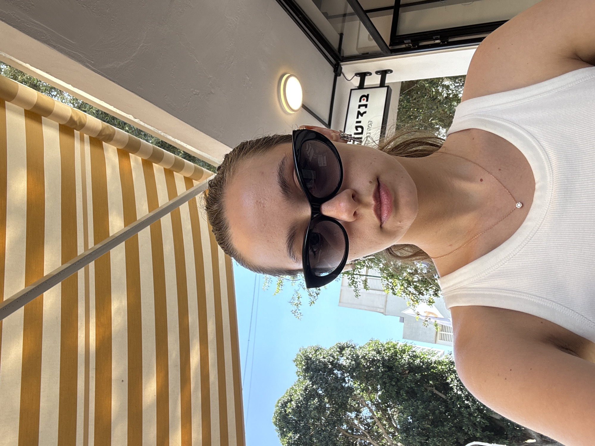 A woman is angled from below, wearing dark sunglasses and a white ribbed tank top with a thin gold necklace. She is sitting under a striped yellow and white awning, likely at an outdoor cafe. The background includes green trees and a clear blue sky. Visible in the upper part of the frame, there's a sign in Hebrew mounted on a black metal frame. Given the location context, the sign is likely for a business in Ramat Gan, Israel. A round light fixture is above the sign. Buildings are seen in the distance amidst the trees.