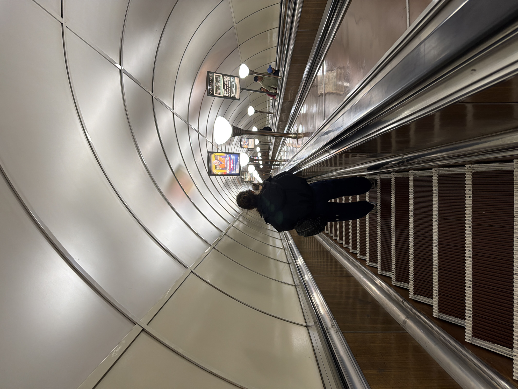 An eye-level, close-up shot inside the Avtovo metro station in Saint Petersburg, Russia, depicts a person standing on a downward-moving escalator. The person is facing away from the camera. Above the escalator, lighting fixtures are interspersed between advertisement boards. The walls lining the tunnel are constructed with a series of curvilinear panels. The escalator itself features metal handrails and dark brown steps. More passengers are visible ahead on the escalator.