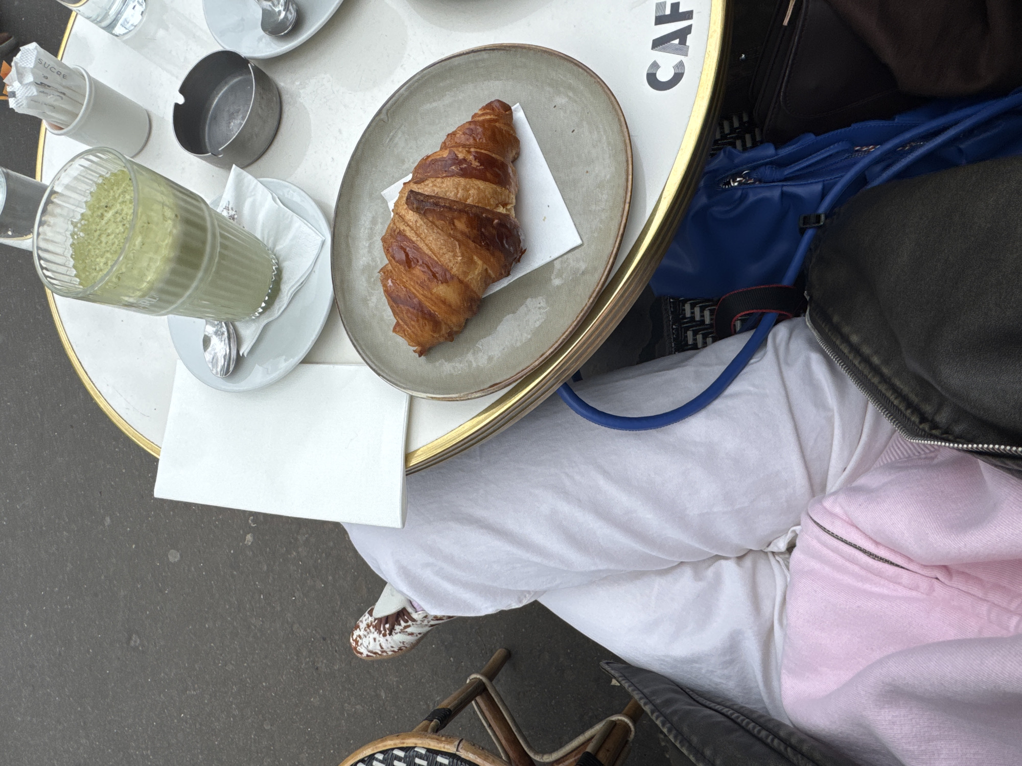 The image shows a table at an outdoor cafe in Paris, France. The table is partially visible with the word "CAFE" printed on it. It holds a plate with a croissant on a paper napkin, a glass of a green-colored beverage, a container of sugar packets, a small dish, and a spoon. A person is seated at the table, wearing white pants, patterned shoes, a pink top, and a jacket. A blue bag and a brown bag are on the chair next to the person. The ground outside the cafe is visible.