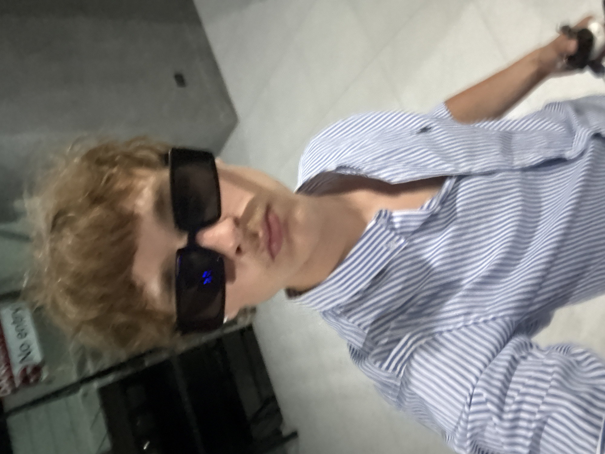 A young male with light brown, curly hair and a light mustache is taking a selfie. He is wearing black, rectangular sunglasses and a blue and white striped button-up shirt. He is standing indoors with a grey wall and floor. To the left, a sign with red lettering and the words "No entry" is visible. The image appears to be taken in Rawai, Thailand.