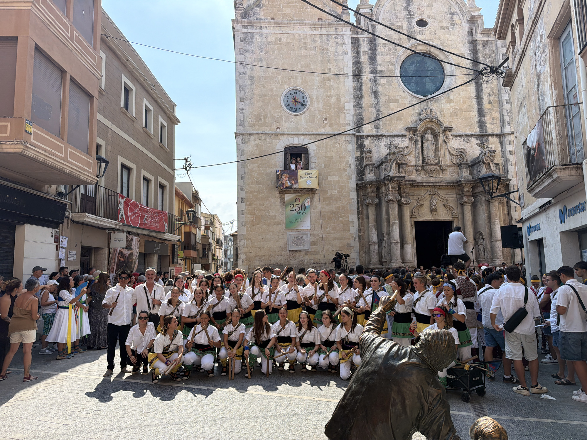 The image depicts a street scene in El Vendrell, Spain, with a large group of people gathered in front of a stone church. Many individuals are dressed in traditional costumes, featuring white shirts, green skirts, and colorful sashes, and some are holding wooden sticks. A statue of a person is visible in the foreground, partially obscuring the view of some of the participants. The church, a prominent architectural structure, features a large clock face on its facade and ornate carvings around its entrance. Several banners and posters are displayed on the church and nearby buildings, suggesting a festive occasion or celebration. The street is paved with cobblestones, and the scene is illuminated by bright daylight, indicating it is daytime. The crowd appears to be engaged in an event, with some individuals posing for photos and others interacting with each other. The presence of a camera on a tripod and the overall atmosphere suggest a public event or performance taking place in the town square.