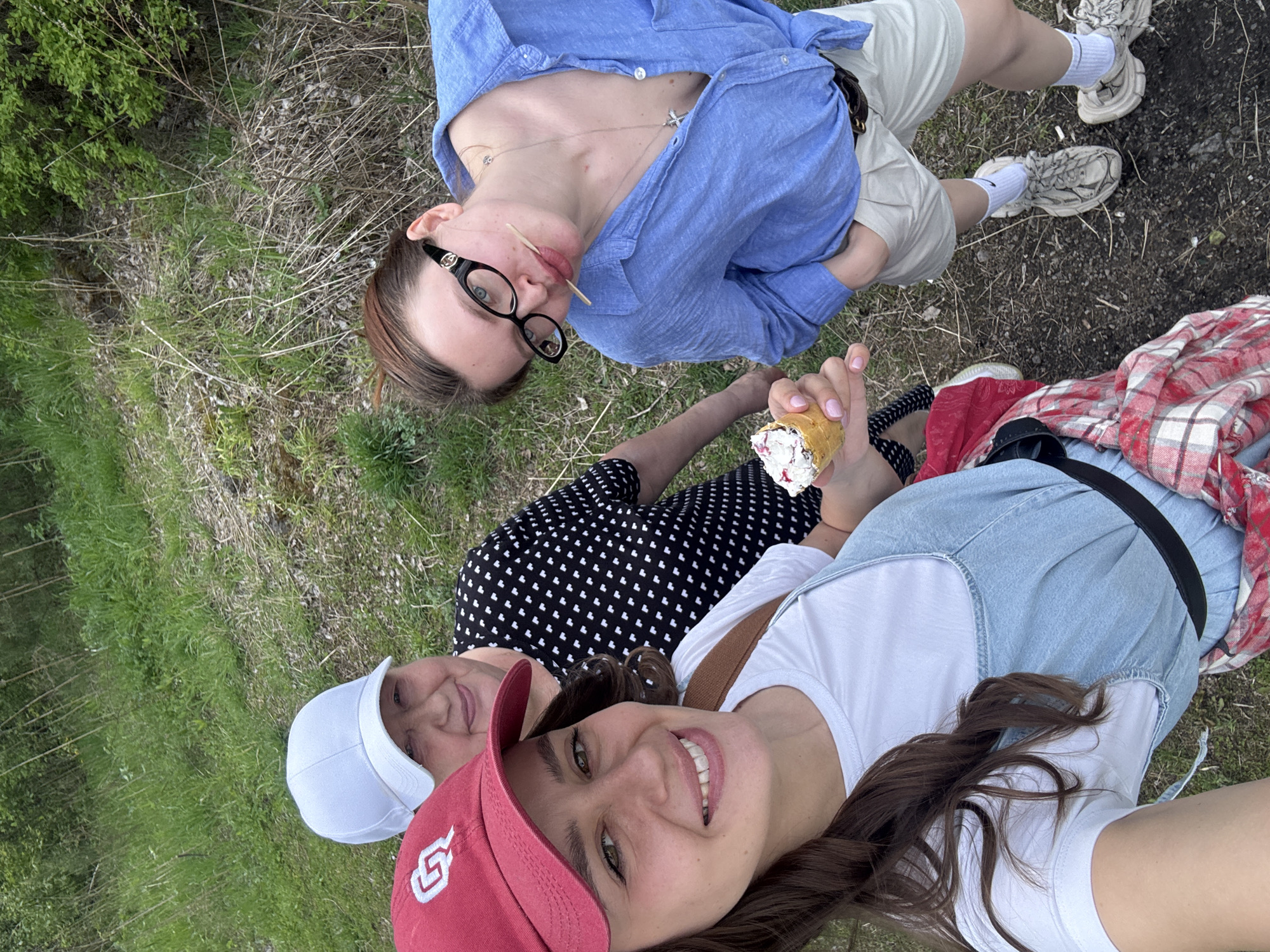 The image shows a group of three people in an outdoor setting that appears to be a grassy area near a road or path. The woman in the foreground wears a red baseball cap and a white t-shirt under denim overalls. She is smiling and taking a selfie. Next to her is a woman in a white baseball cap and a black and white patterned dress. Further back, a third woman wearing a blue button-down shirt, beige shorts, and glasses is looking into the camera with two toothpicks in her mouth. In her left hand, another woman holds an ice cream cone. The landscape includes grass, some dried vegetation, and a paved surface on the right side. Based on information available, it is not possible to determine the exact location as the city name does not appear in the image.