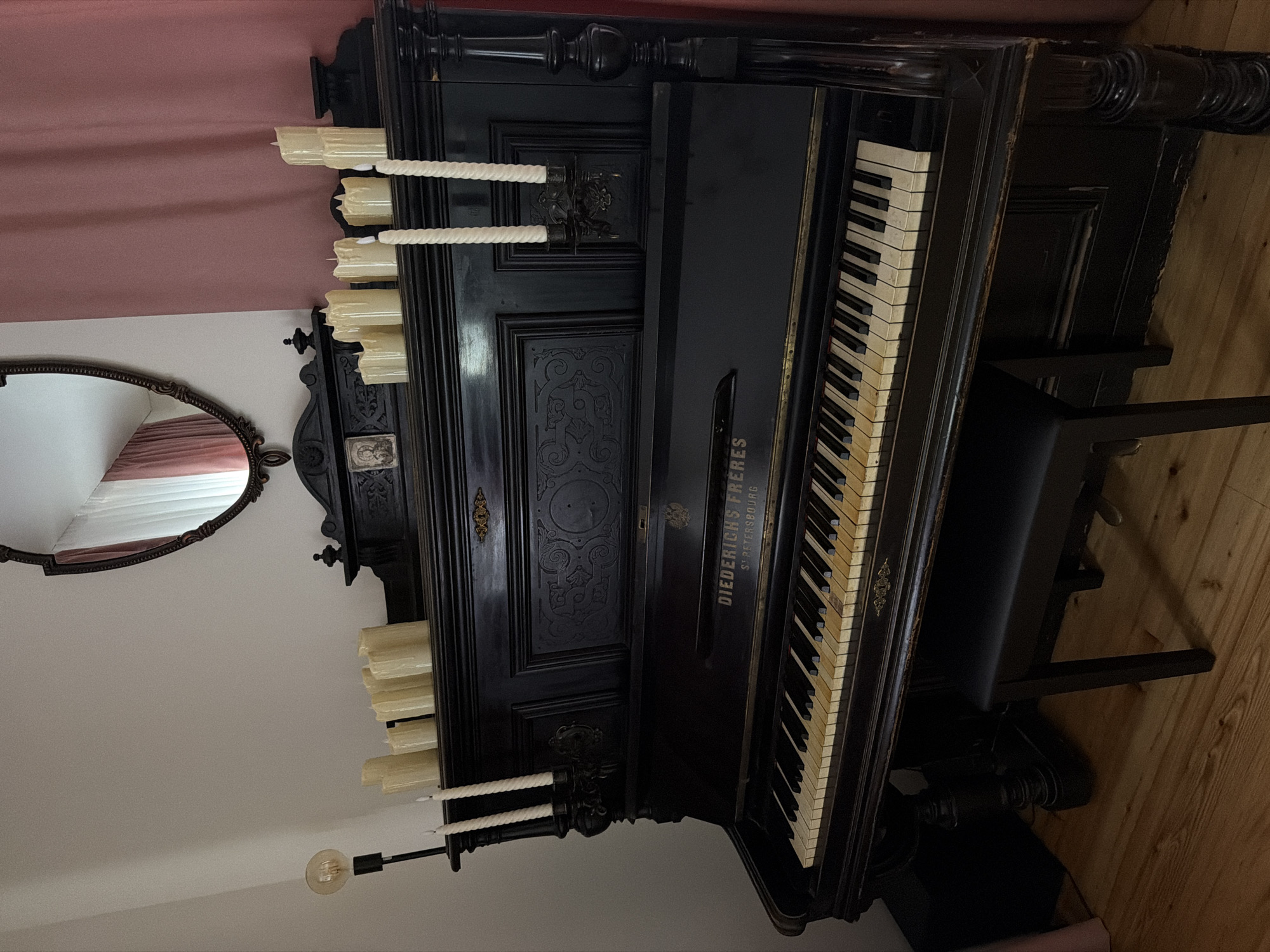 An antique black upright piano with yellowed keys and decorative carvings sits on a wooden floor. A black piano bench is positioned in front of it. Several tall, unlit pillar candles are placed on the top of the piano, with two more candles in a candelabra on the right side. A lightbulb on a black stand is to the left of the piano. Behind the piano, a dark wooden mirror with an ornate frame is hung on a light-colored wall, reflecting a pink curtain. The scene appears to be indoors, possibly in a room with a warm, vintage ambiance. There is no specific indication of the location being in Kolomna, Russia.