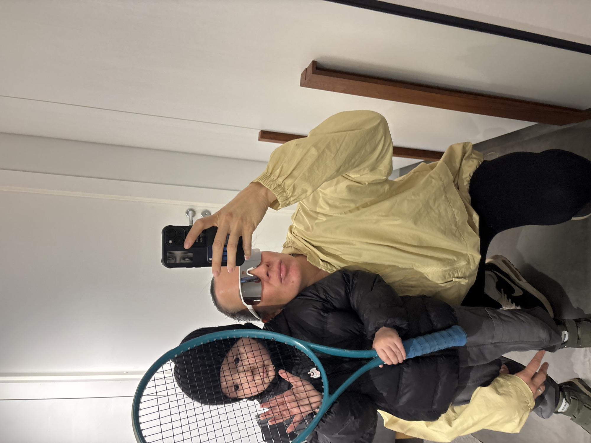 An adult and a child are captured in a mirror selfie indoors. The adult, positioned behind and slightly to the side of the child, is wearing a light yellow or cream-colored long-sleeved jacket and distinctive white-framed, reflective sunglasses. They hold a black smartphone with a prominent camera array and a dark case, angled to take the photo.
The child stands in the foreground, wearing a black puffer jacket, a dark beanie, and gray pants. They are holding a blue-framed tennis racket with a blue grip, looking directly at the camera through the racket's strings. Their face shows a neutral expression.
The adult's footwear, partially visible, includes a sneaker with a white sole and a dark upper, featuring a clear "Nike" logo. The child wears green shoes with hook-and-loop fasteners. The background consists of light-colored walls or panels, possibly white, with horizontal dark lines and some wooden trim elements visible higher up, suggesting a modern interior space like a hallway or changing room. No specific time of day or weather is evident. No other text beyond the "Nike" logo is clearly legible within the image.