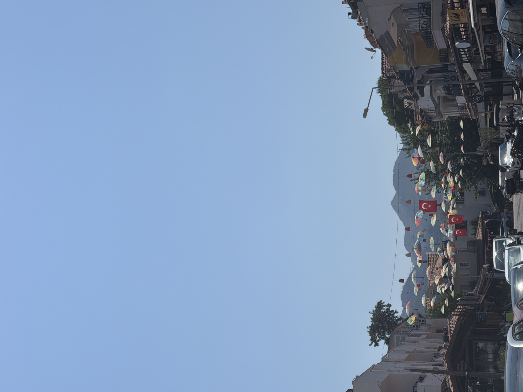 The image depicts a street scene in Marmaris, Turkey, decorated with numerous colorful umbrellas and Turkish flags suspended from overhead wires. The umbrellas are of various colors and patterns, creating a vibrant canopy above the street. The Turkish flags, featuring a white star and crescent on a red background, are interspersed with the umbrellas.
The street itself is lined with buildings, some of which appear to be shops or restaurants, with awnings and outdoor seating visible. Cars are parked along the street, and a few motorcycles are also present. In the background, mountains are visible under a clear blue sky. The architecture suggests a Mediterranean or coastal town setting. The overall impression is that of a lively and festive street, likely a popular area for tourists or locals. There are no prominent individuals or specific actions captured in the frame, making it a general scene of a decorated street.