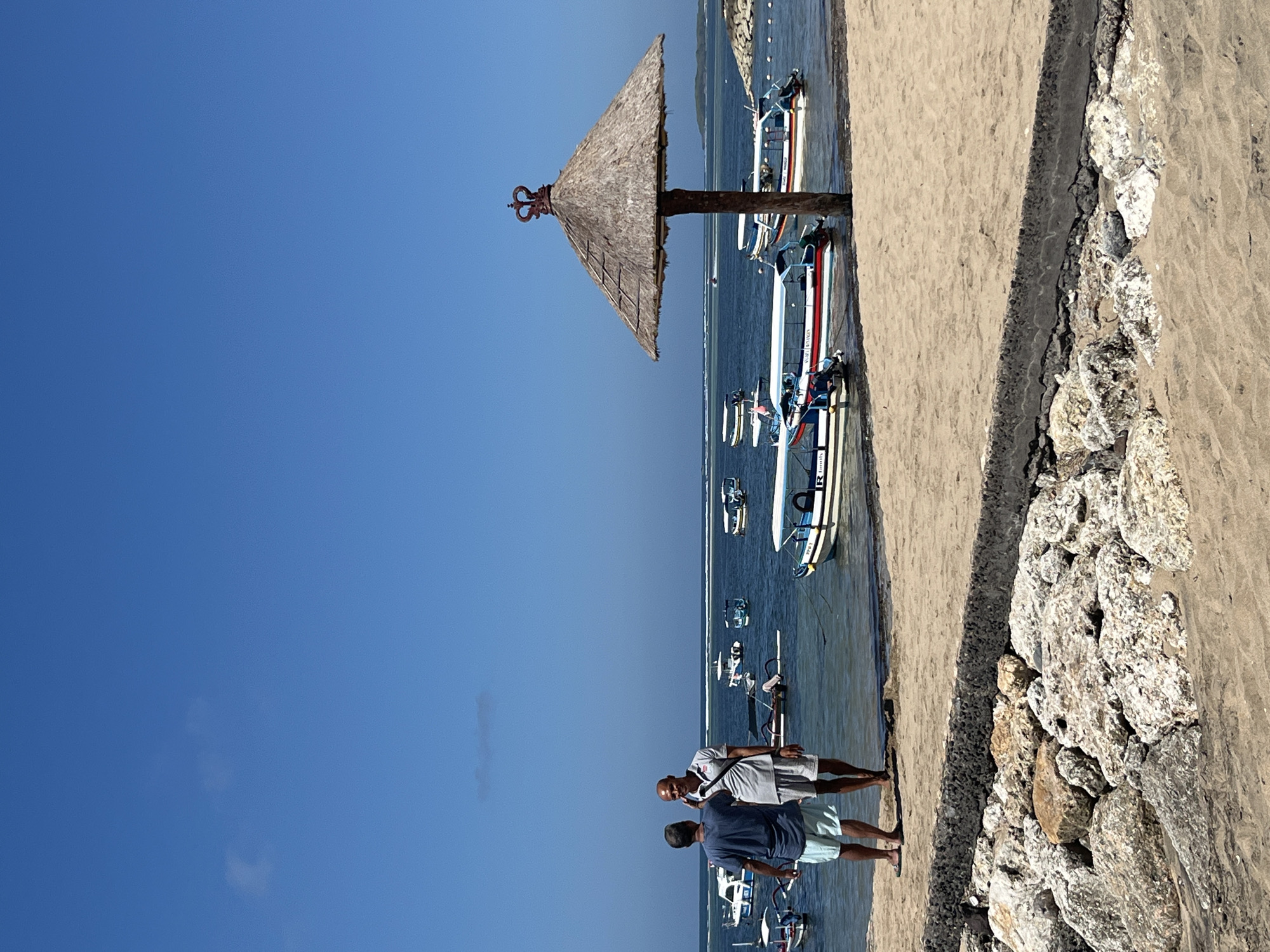 The image depicts a serene beach scene in Kuta Selatan, Indonesia, under a clear blue sky. In the foreground, a sandy beach transitions into a rocky embankment, suggesting a man-made barrier. Two men are standing near the water's edge, engaged in conversation. One man, wearing a blue t-shirt and light-colored shorts, stands with his back to the camera, while the other, in a grey polo shirt and shorts, faces him.
The water is calm and dotted with several boats, likely tourist vessels, including some with thatched roofs. A prominent thatched umbrella stands on the sand, providing shade. In the distance, a thin line of the horizon separates the sea from the sky, with a faint suggestion of landmasses. The overall impression is one of a tranquil tropical environment, possibly a tourist destination given the presence of boats and the beach umbrella.