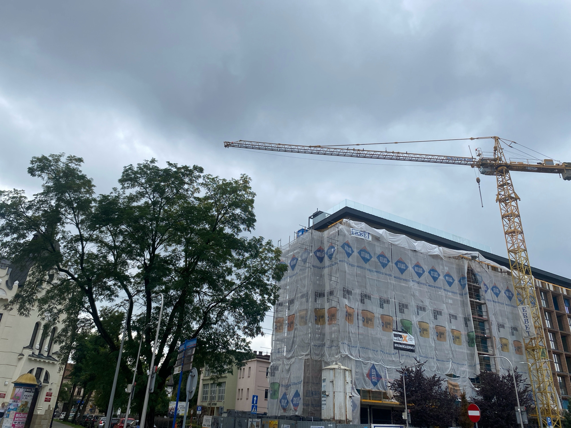 The image shows a construction site in Nowy Sącz, Poland, with a yellow tower crane extending over a building covered in white scaffolding netting. The netting displays various logos and images, including those of "ATLAS" and "GEMINI RS". A portion of a dark-colored building with a modern glass railing is visible at the top of the construction site. To the left of the construction site, a large tree with dense green foliage stands next to an older, light-colored building with arched windows. Street signs and lampposts are also present on the street in front of the buildings. The sky is overcast with gray clouds.