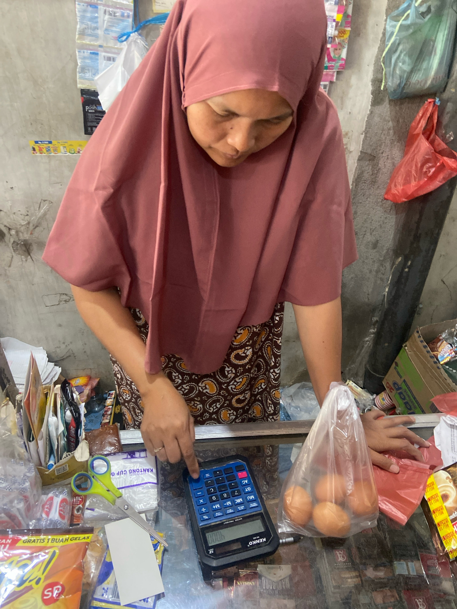 In Kaliwungu, Indonesia, a woman wearing a dusty rose headscarf and a patterned brown and orange skirt stands behind a glass counter. She is looking down at a blue Kenko calculator, her right index finger pressing a button on its keypad. To her left, a pair of green-handled scissors lies on top of various packaged goods, including what appears to be food items and drinks. A small white card sits near the scissors. To her right, a clear plastic bag contains four brown eggs. The counter is cluttered with more packaged products and a red plastic bag. In the background, the wall is rough and appears to be made of concrete blocks, with various items hanging on it, including plastic bags and colorful packaging. A cardboard box filled with more products is visible to the right. The overall scene suggests a small shop or market stall.