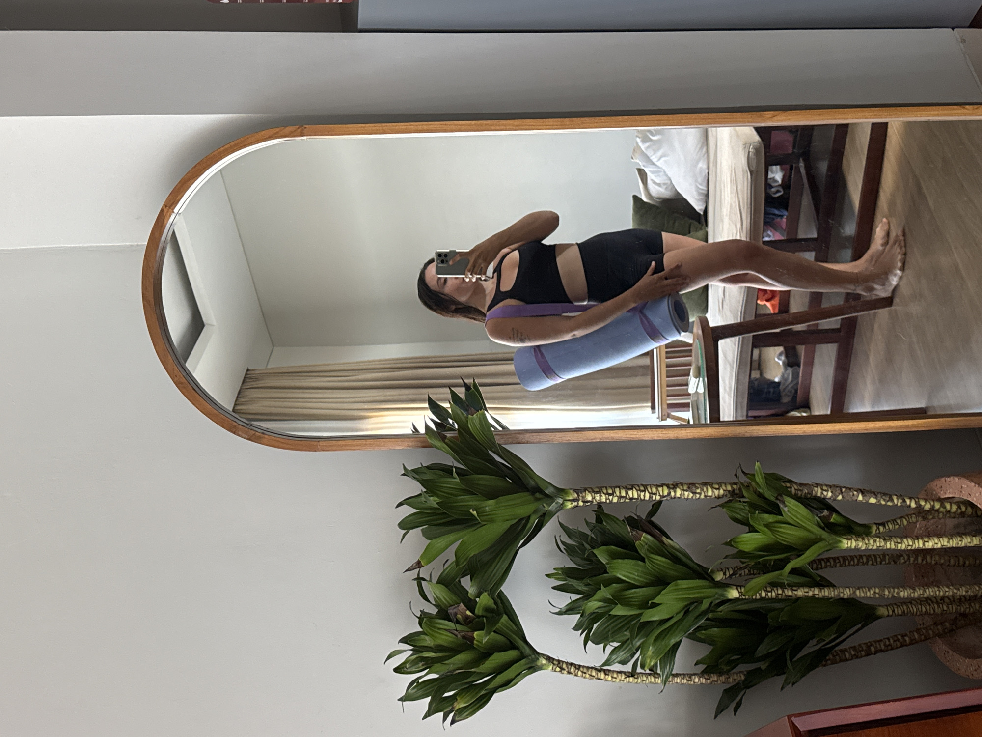 A woman stands in front of a full-length mirror, taking a selfie. She is wearing a black sports bra and black shorts and holding a rolled-up blue yoga mat with a purple strap over her shoulder. The mirror is framed in wood and has an arched top. To the left of the mirror are three tall green plants in a terracotta pot. In the reflection of the mirror, a bed with white and green pillows can be seen, along with a wooden table and chair. The scene appears to be indoors, possibly a bedroom or a living area, in a room with light-colored walls and sheer curtains in the background. The floor is wooden. The woman's pose and the presence of the yoga mat suggest she is about to engage in or has just finished a yoga session. The overall impression is one of casual fitness and a relaxed home environment.
