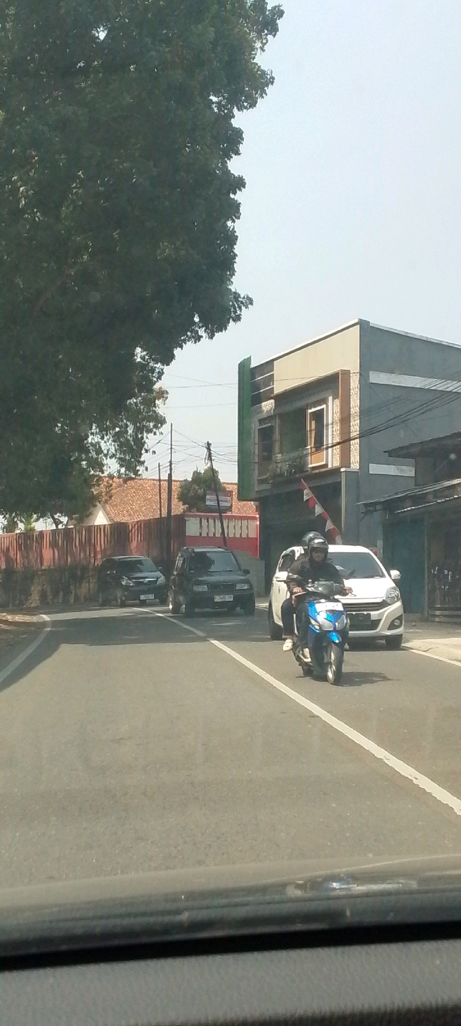 In Susukan, Indonesia, on a sunny day, a motorcycle with two occupants, a male driver wearing a helmet and a female passenger, travels down a paved road. The motorcycle is blue and white. Behind them, a white car is visible, and further down the road, two dark-colored SUVs are also present. The scene is lined with trees on the left and buildings on the right. The road has white lane markings. A portion of a dashboard is visible in the foreground of the image.