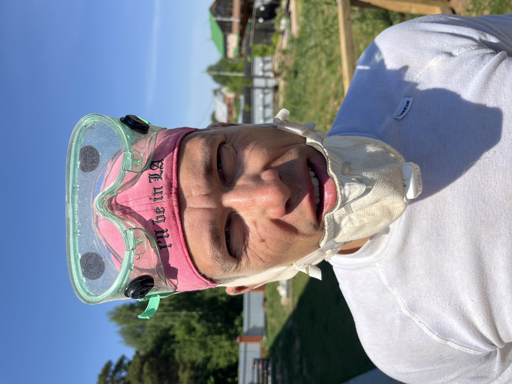 A person, presumably male, is seen in close-up, wearing protective gear. This includes translucent green goggles, a pink head covering, and a white facial mask. The person is wearing a white t-shirt. The background shows a sunny day with green vegetation, a wooden fence, and buildings in the distance. The location is the city of Chaikovsky, Russia.