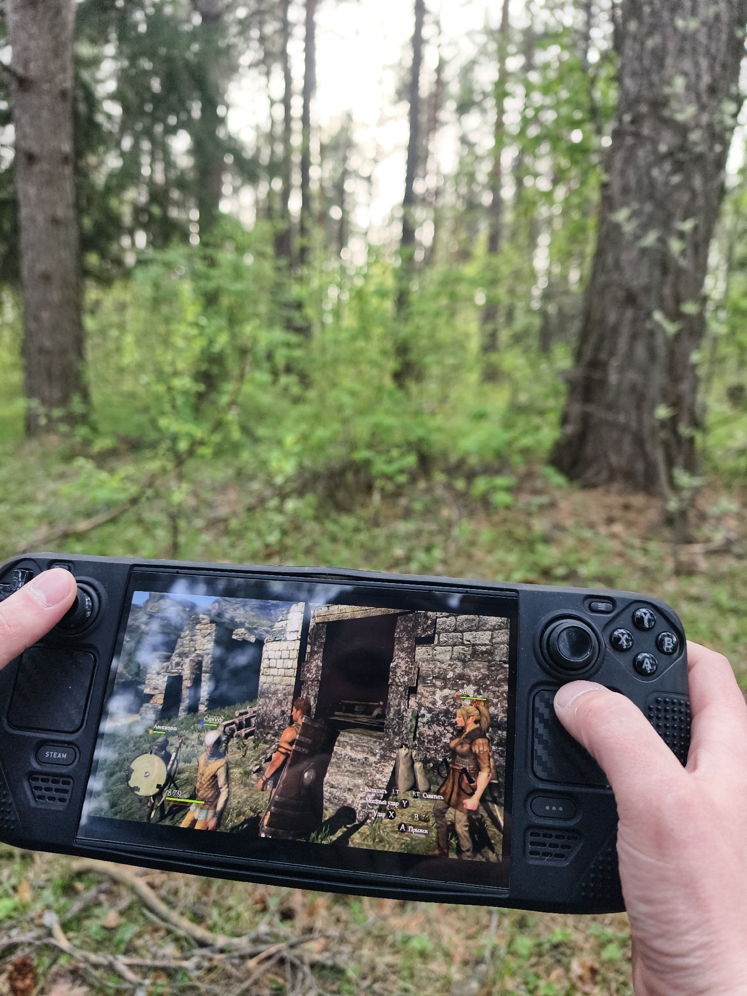 A person is holding a black Steam Deck gaming console, displaying a video game. The game appears to be set in a medieval or fantasy environment, featuring characters in rustic clothing standing in front of a stone structure. The user is using the right-hand controls of the Steam Deck. In the background, out of focus, is a natural forest setting, likely in the city of Прикамский, Russia.