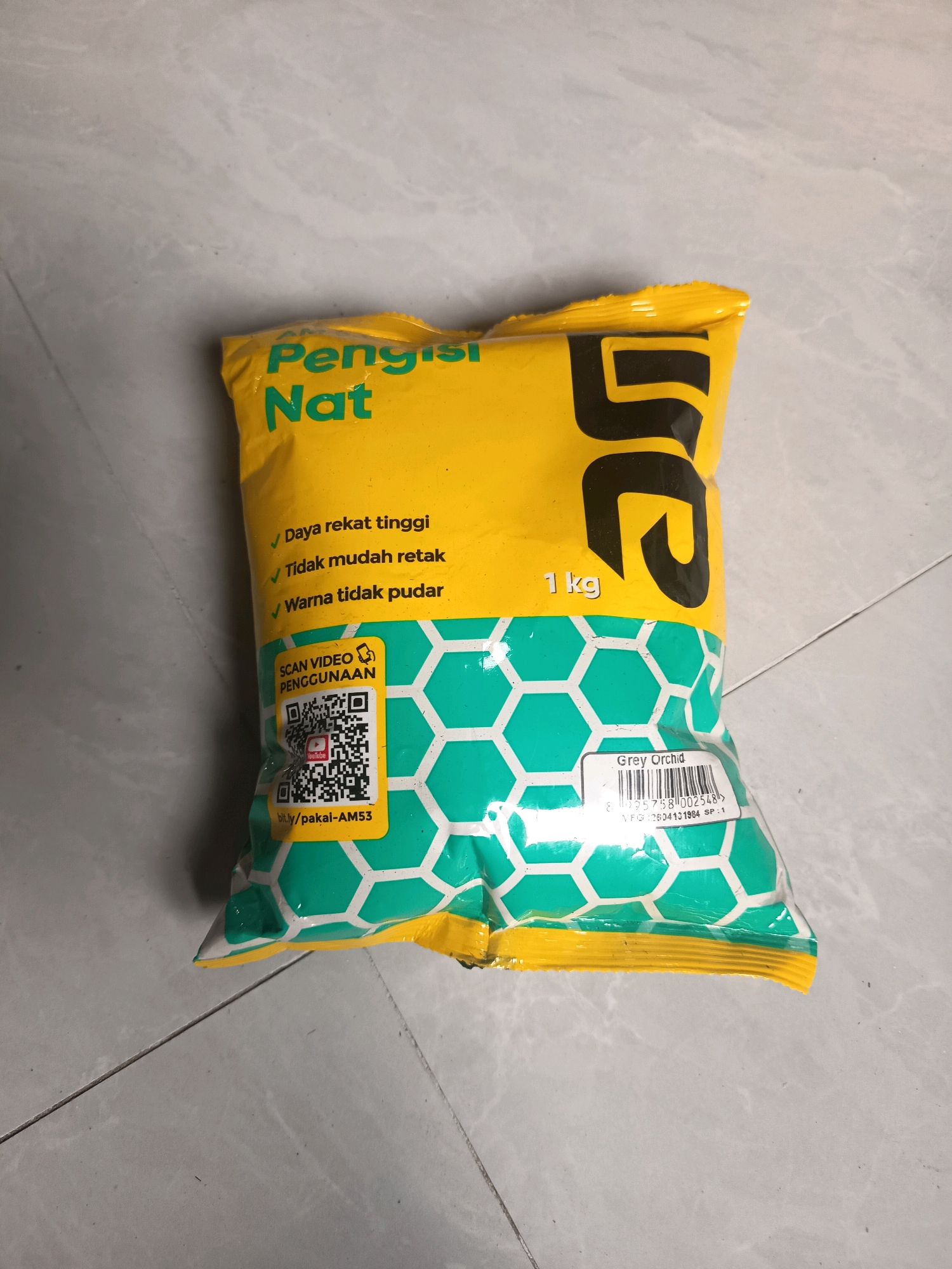 A yellow and green package of "Pengisi Nat" is lying on a grey tiled floor. The package is oriented vertically, with the top of the package facing the top of the image. The package has text in Indonesian, including "Pengisi Nat," "Daya rekat tinggi," "Tidak mudah retak," and "Warna tidak pudar." There is also a QR code labeled "SCAN VIDEO PENGGUNAAN" and a URL "bit.ly/pakai-AM53." A barcode and the text "Grey Orchid" are also visible on the package. The package is approximately 1 kg in weight. The floor appears to be made of light grey tiles with dark grey grout lines. The lighting suggests the photo was taken indoors, likely in a residential or commercial space. No people are visible in the image. The setting is a generic tiled surface, not specifically indicative of any particular location within Sidorejo, Indonesia, or any other city.