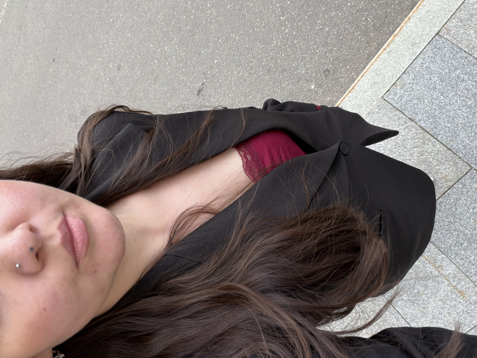 A close-up, overhead shot shows a person's face and upper body, rotated 90 degrees counter-clockwise. The person has long, dark brown hair that falls across their shoulders and a black blazer. A sliver of dark red lace, likely from lingerie, is visible beneath the open blazer. The person's face is partially visible, with closed eyes, light pink lips, and a nose piercing. The background is an outdoor setting with textured gray pavement and lighter gray stone tiles. The lighting suggests it is daytime, possibly overcast, as there are no strong shadows. The overall mood is casual and relaxed.