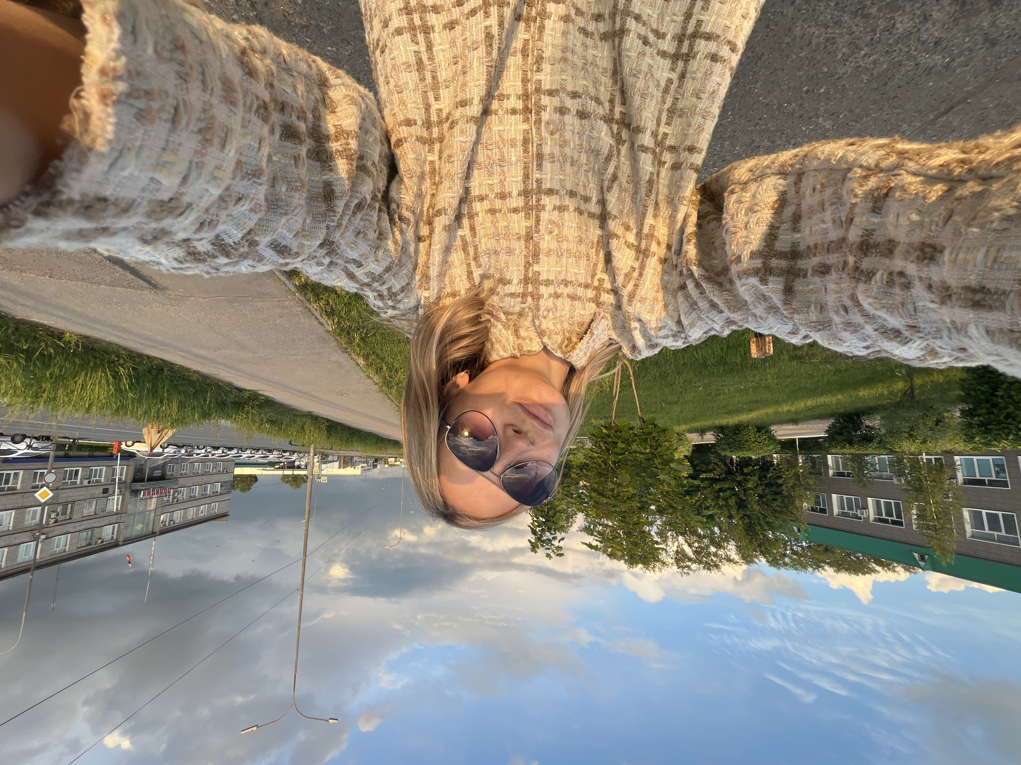 A vertically oriented, low-angle selfie photograph presents a woman outdoors, likely in the Russian city of Chaikovsky. She is fair-skinned with light blonde hair and wears sunglasses, posing in a plaid, tweed-like jacket or coat. The background reveals an urban scene, encompassing a paved road adjacent to a grassy verge, buildings, and a sky filled with cumulus clouds. One building bears signage in Cyrillic, identifying it as a police station. The photograph's composition emphasizes the woman and the surrounding urban environment.