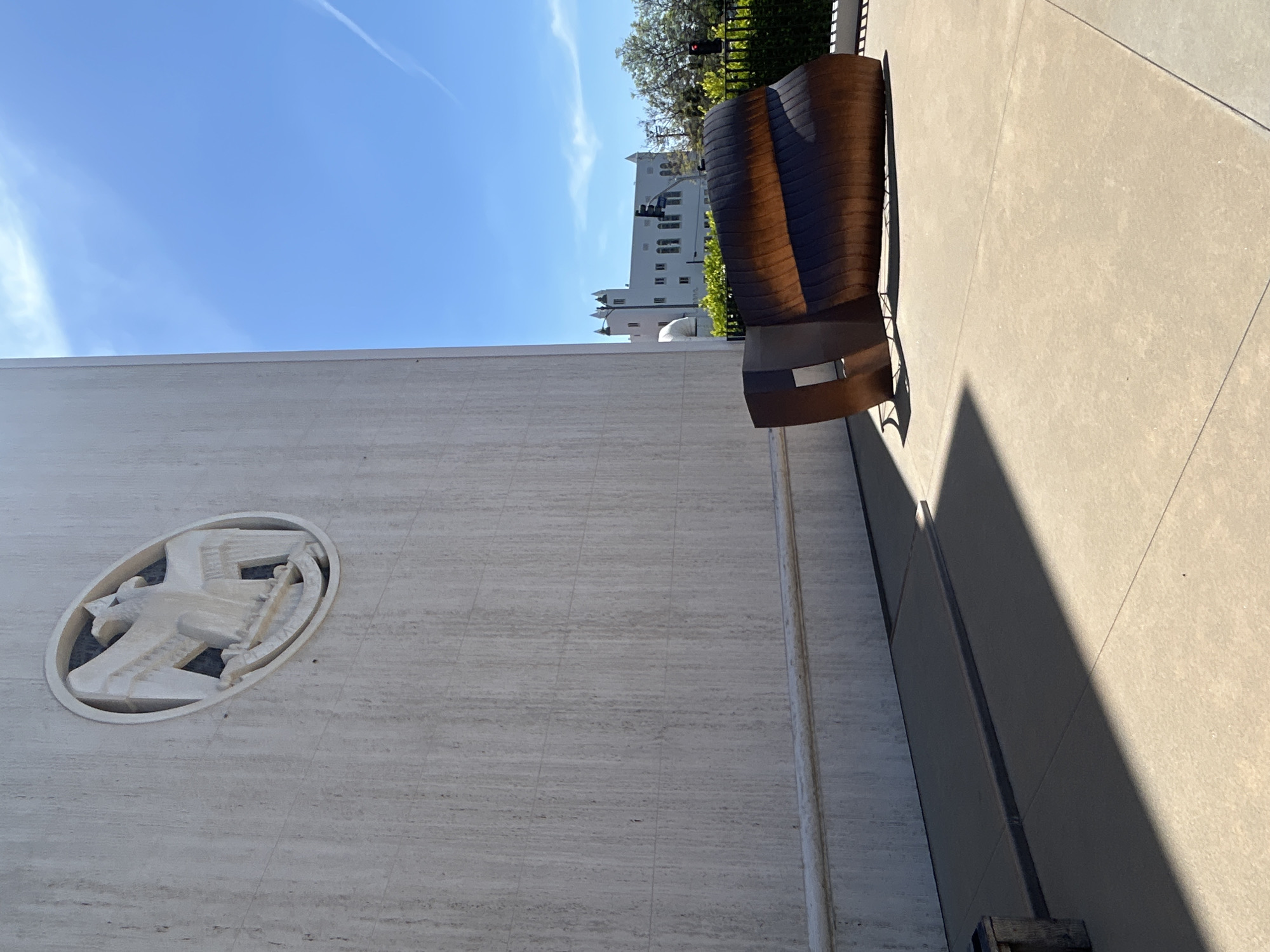 The image shows a view of the Los Angeles Central Library in Los Angeles, California, United States of America. The focus is on the library's exterior wall, which features an oval-shaped carved emblem of a stylized bird holding a book. Next to the library's wall, there is a large, abstract sculpture which looks like a chair. The background includes a blue sky and a building with multiple windows and spires. The scene is well-lit with natural sunlight, casting a shadow on the sidewalk next to the building.