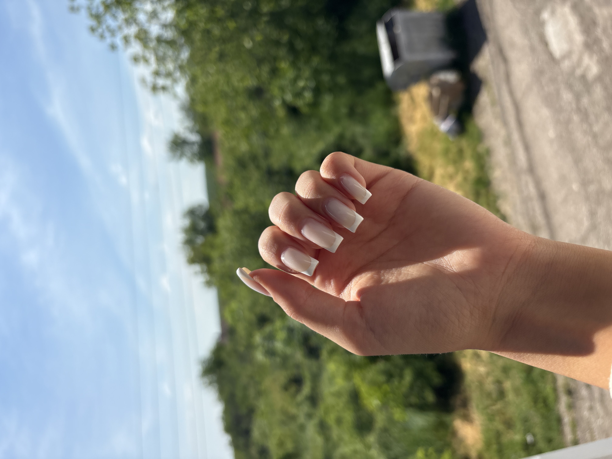 The image shows a hand with long, white, square-tipped nails. The hand is fair-skinned and positioned against a blurred background of green foliage and a partly visible blue sky. The background appears to be outdoors, with trees and possibly other vegetation visible. The top right corner shows a section of concrete. There is insufficient information to determine the location in Medkovets, Bulgaria with certainty.