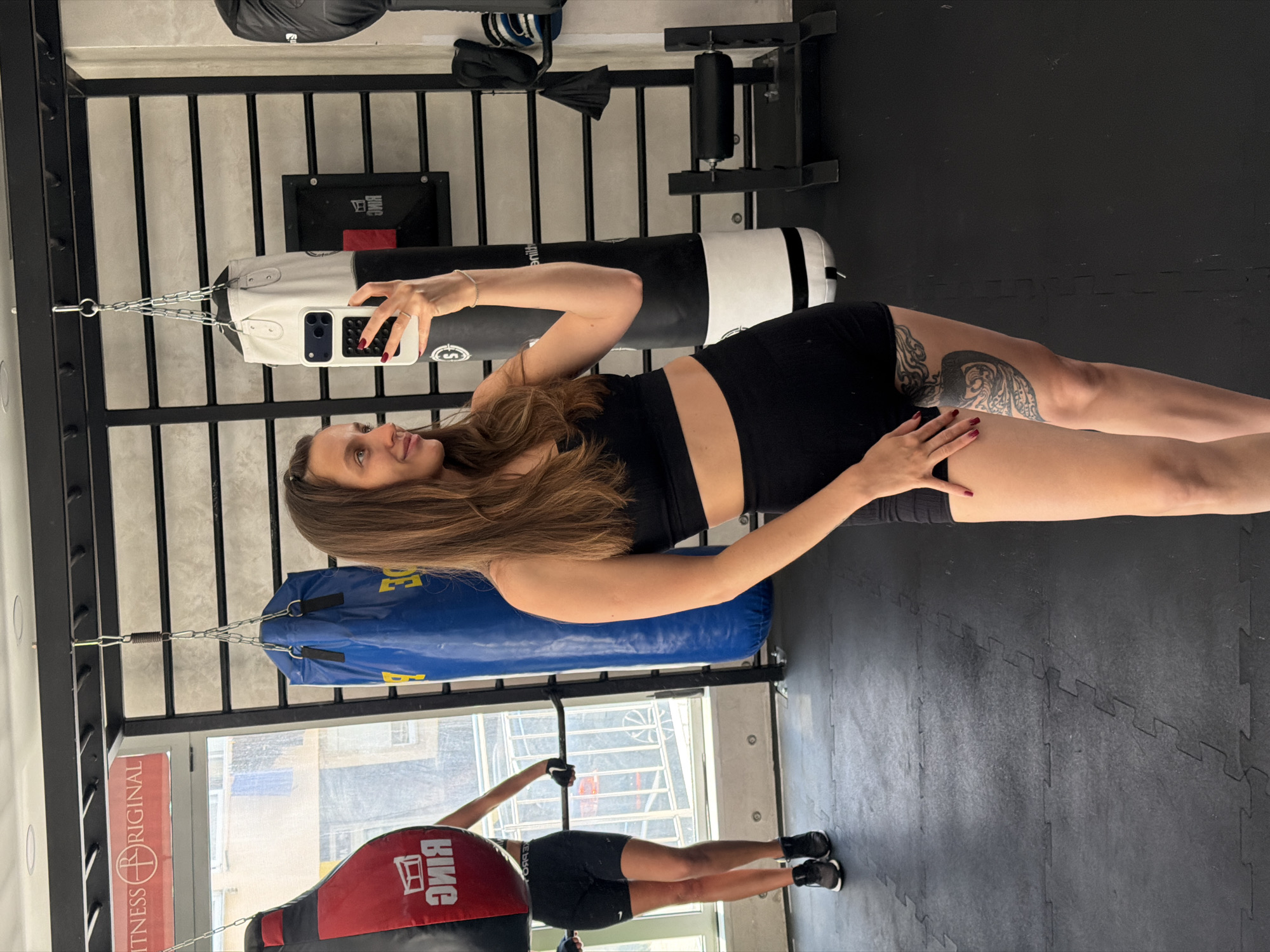 A woman is standing in a gym in Budva, Montenegro, facing a window. She is wearing a black sports bra and shorts. She is taking a selfie with a white phone, and there is a red and blue punching bag hanging in the background. Another person is standing in front of the window, also in athletic clothing.