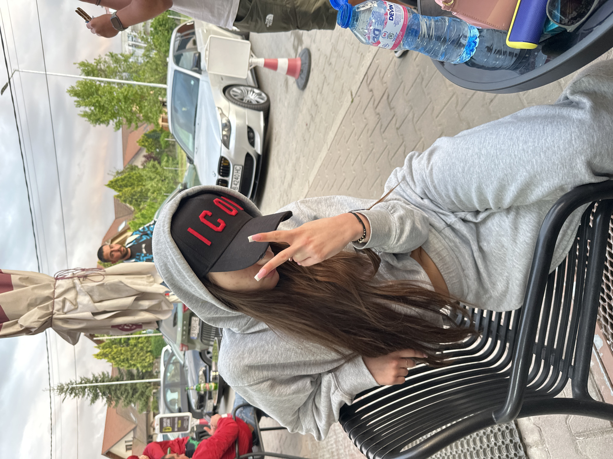 The image shows a woman sitting at an outdoor cafe, wearing a grey hoodie and a black baseball cap with the word "ICON" in red lettering. She is making a peace sign with her hand. A water bottle, a wallet, and sunglasses are on the table. In the background, there are other people, cars, and buildings. The location is likely in Plovdiv, Bulgaria.