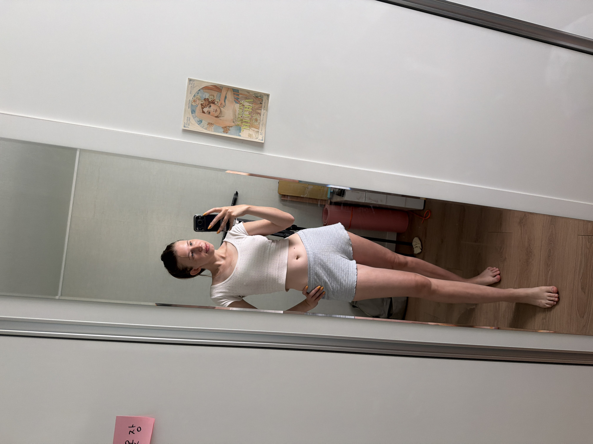A young woman with her hair in a bun takes a mirror selfie in what appears to be a bedroom. She is wearing a white cropped t-shirt and grey shorts. Her left hand is on her hip and she is holding a smartphone in her right hand. The room has a wooden floor and a white wardrobe. A pink sticky note with Korean writing is affixed to the wardrobe. A framed print of a woman is also on the wardrobe. The image was taken in Dongducheon-si, South Korea.