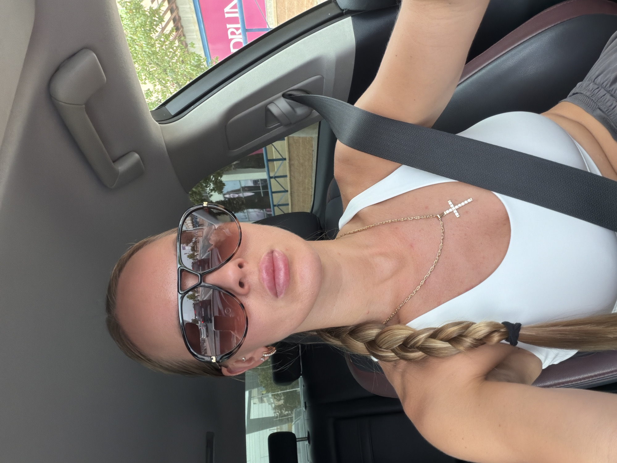 A blonde woman with a braid is sitting in the driver's seat of a car, wearing oversized sunglasses, a white sports bra, and grey sweatpants. She is wearing a gold chain with a diamond-encrusted cross pendant. The interior of the car is visible, including a seatbelt and a ceiling handle. The woman's sunglasses reflect the road ahead, showing other cars and a building with pink signage that appears to say "ORLIN". The background suggests an urban or suburban environment. Based on the signage, the location is identified as Finestrat, Spain.