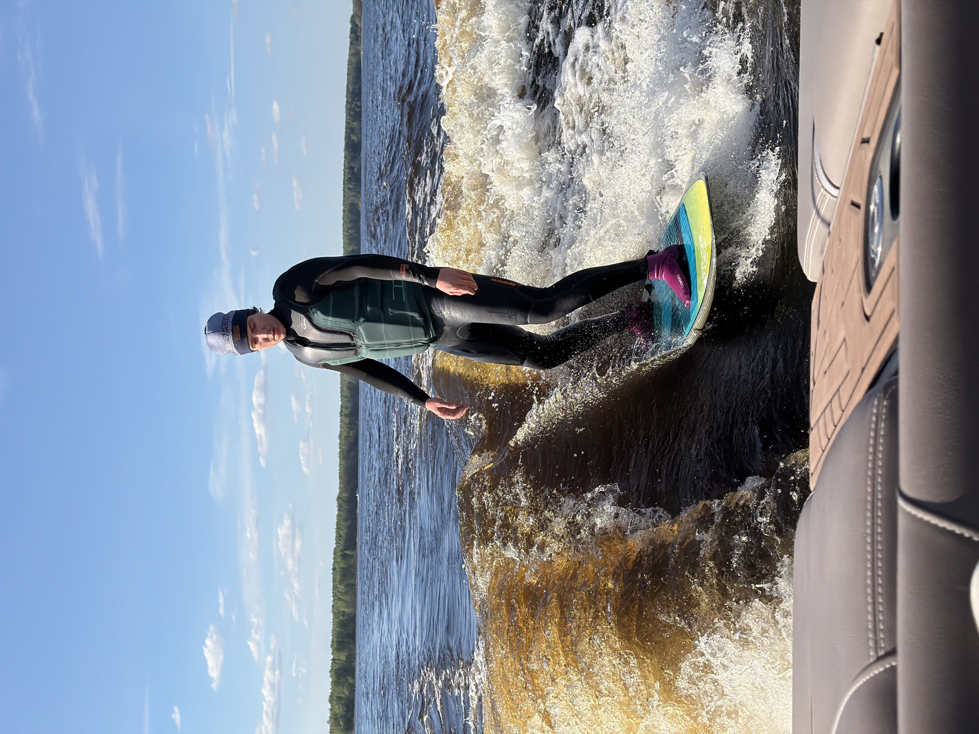 A person is wake surfing on a lake, likely in Gorodishche, Russia, as indicated by the prompt. The person is wearing a black wetsuit, a light green life vest, and a blue cap, and is riding a yellow and blue wakeboard with pink boots. They are positioned to the right of the frame, riding the wake of a boat. A portion of the boat's interior is visible on the right side of the image. The lake appears to be surrounded by a forest in the background, and the sky is blue with scattered clouds. The water is choppy, with a mix of dark blue and foamy white where the wake is.