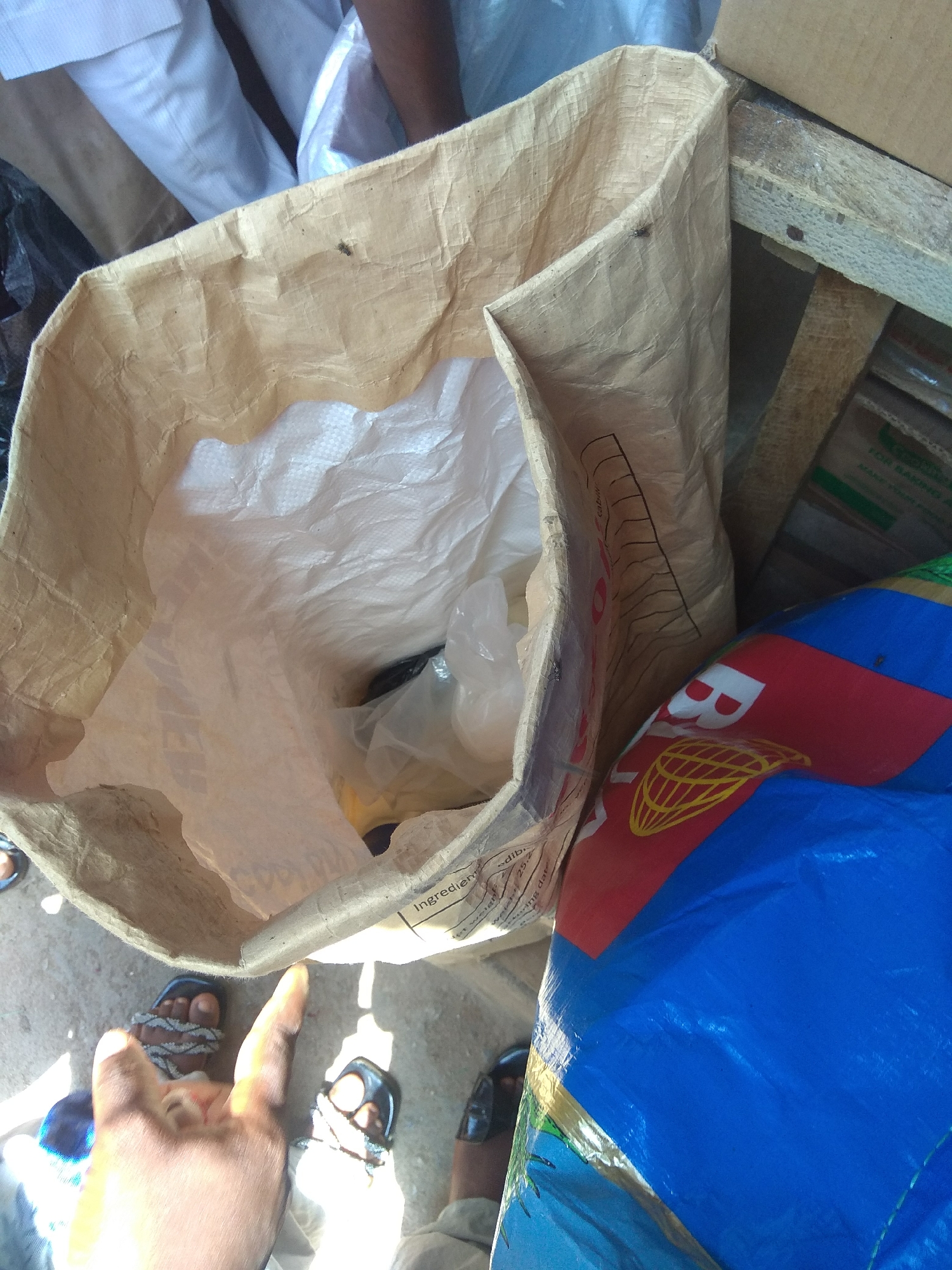 The image depicts an outdoor scene in Jalingo, Nigeria, likely a market or a storage area. In the foreground, a large, open, tan-colored woven bag made of a synthetic material is prominently displayed, with its top edges rolled back. The interior of the bag is lined with a white, crinkled material, possibly plastic or another protective layer, and appears to contain some objects wrapped in clear plastic.
To the right of the large bag, a portion of a blue, crinkled plastic material with red and yellow markings is visible. The markings include a globe emblem with yellow lines forming a grid over it and parts of white letters. This object appears to be another bag or cover.
In the lower section of the image, parts of people's feet are visible. One person is wearing sandals with decorative straps. Another set of feet is wearing black sandals. The ground is a dusty, concrete surface, suggesting an outdoor, possibly unpaved, area. There are also some wooden planks visible, suggesting a makeshift structure or pallets. In the background, the legs of people wearing white clothing are discernible, indicating human presence in the vicinity. The overall impression is one of commerce or goods being handled in an open, possibly busy, environment.