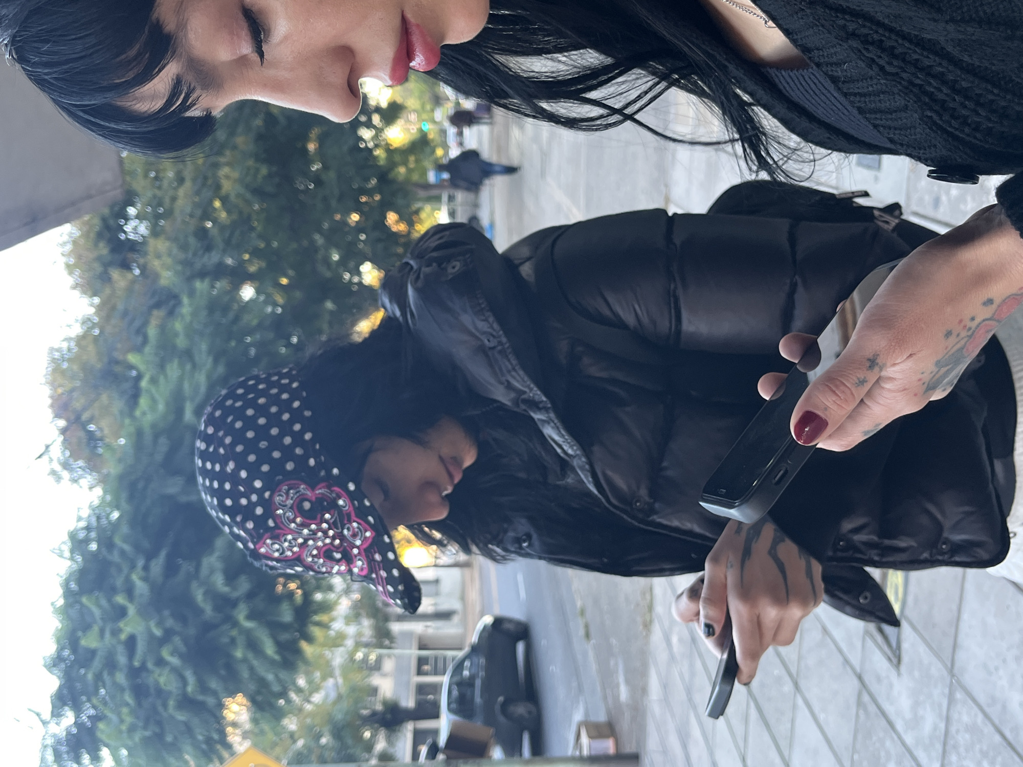 Two young women are standing on a sidewalk in Buenos Aires, Argentina, looking at smartphones. The woman closest to the camera has long black hair, fair skin, and red lipstick. She is wearing a black sweater. The other woman is wearing a black polka-dotted bonnet and a black puffer jacket. She has tattoos on her hands and arms. A street with cars is visible in the background. Trees are also visible.